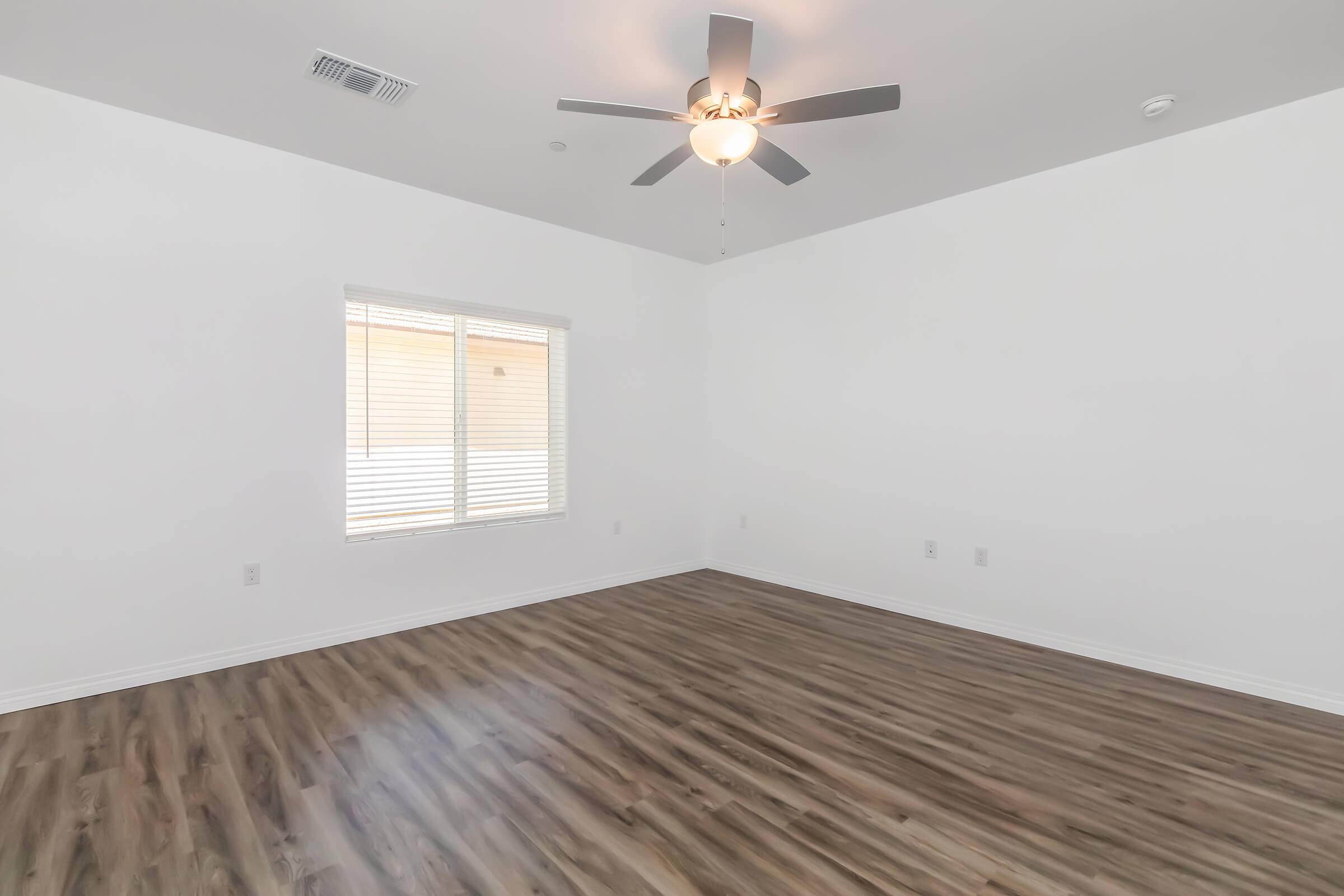 A bright, empty room with light-colored walls and a wood laminate floor. It features a ceiling fan and a window with closed blinds, allowing natural light to enter. The space is clear of furniture, providing a sense of openness.