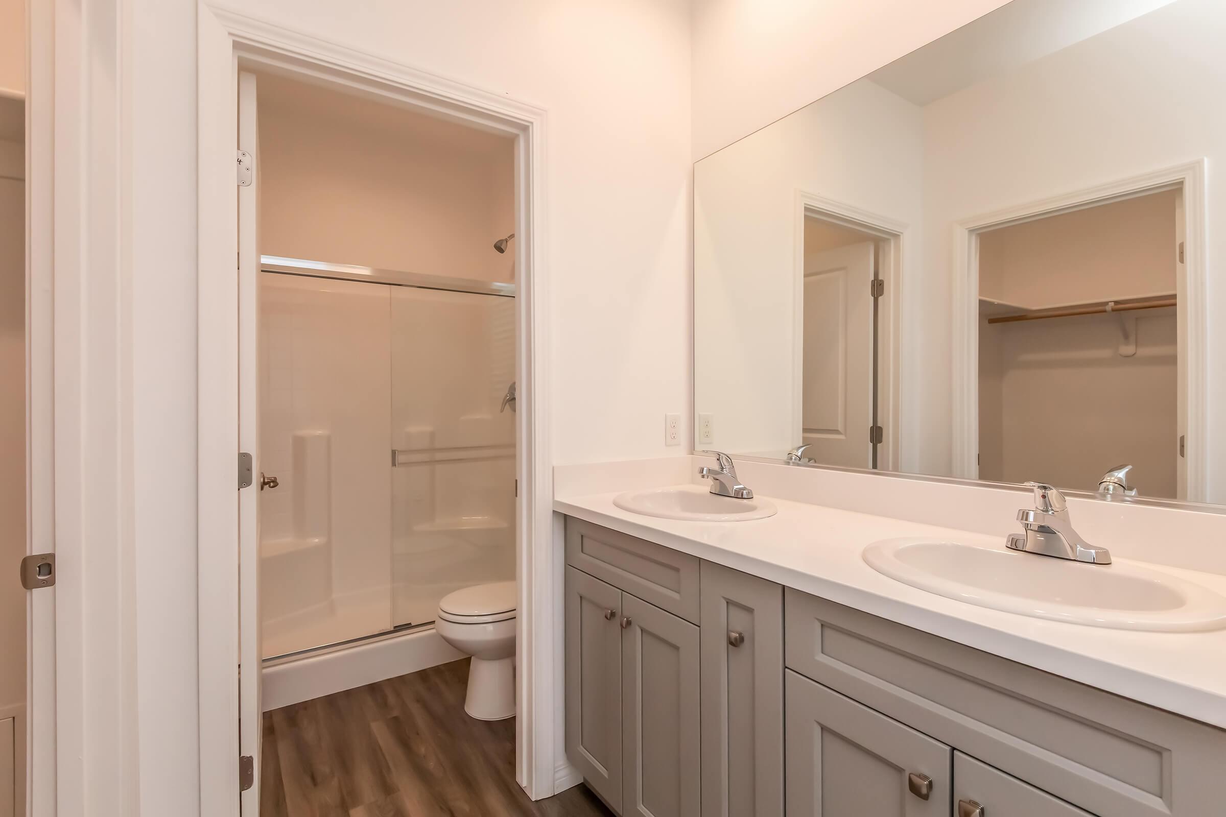A modern bathroom featuring a double vanity with two sinks and mirrors, a spacious shower with glass doors, and a toilet. The room has light neutral walls and a wood-like floor, creating a clean and contemporary aesthetic. A closet is visible in the background.