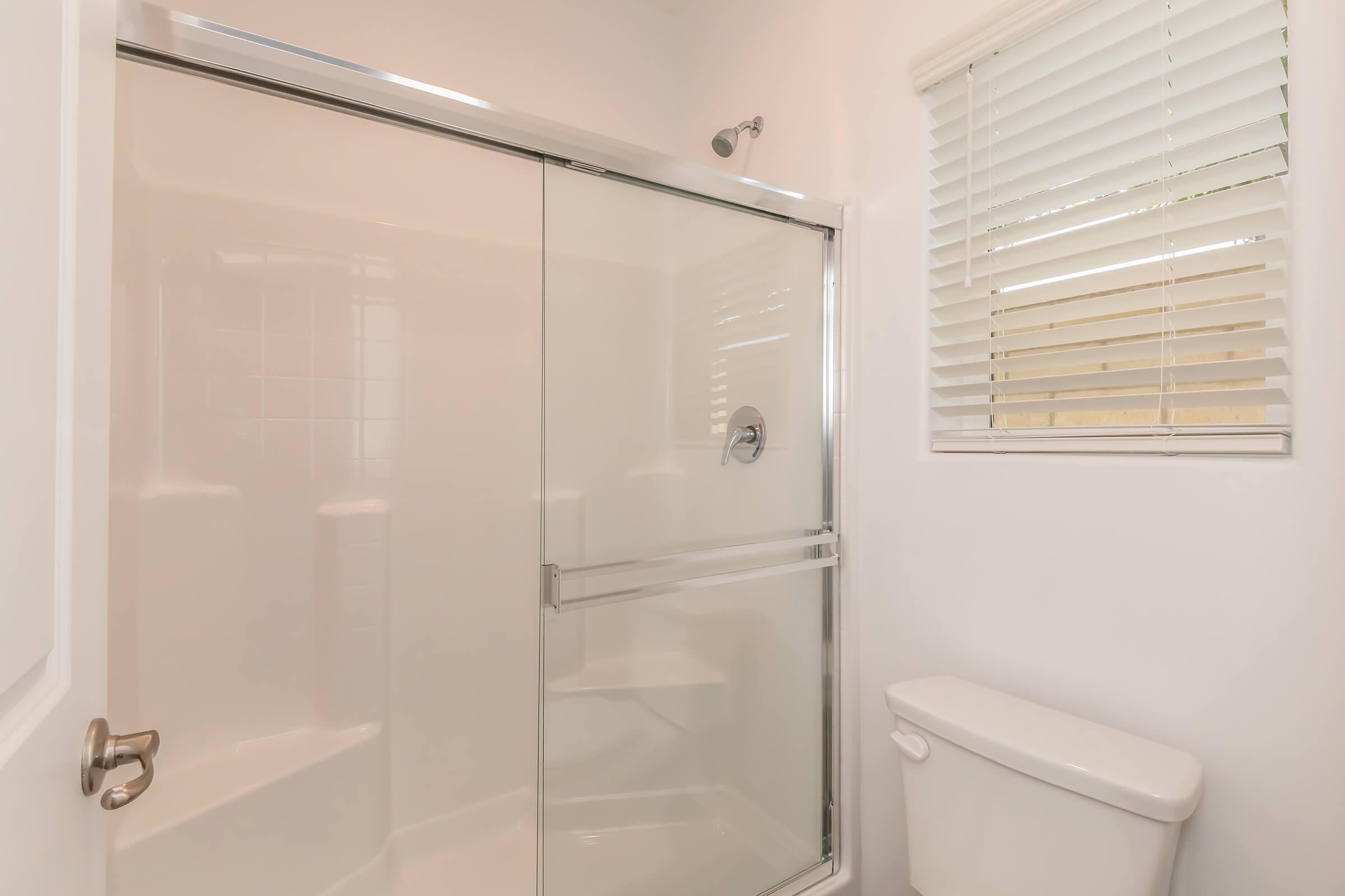 A clean and modern bathroom featuring a glass shower enclosure, a simple white toilet, and a window with horizontal blinds letting in natural light. The walls are painted white, creating a bright and airy ambiance.