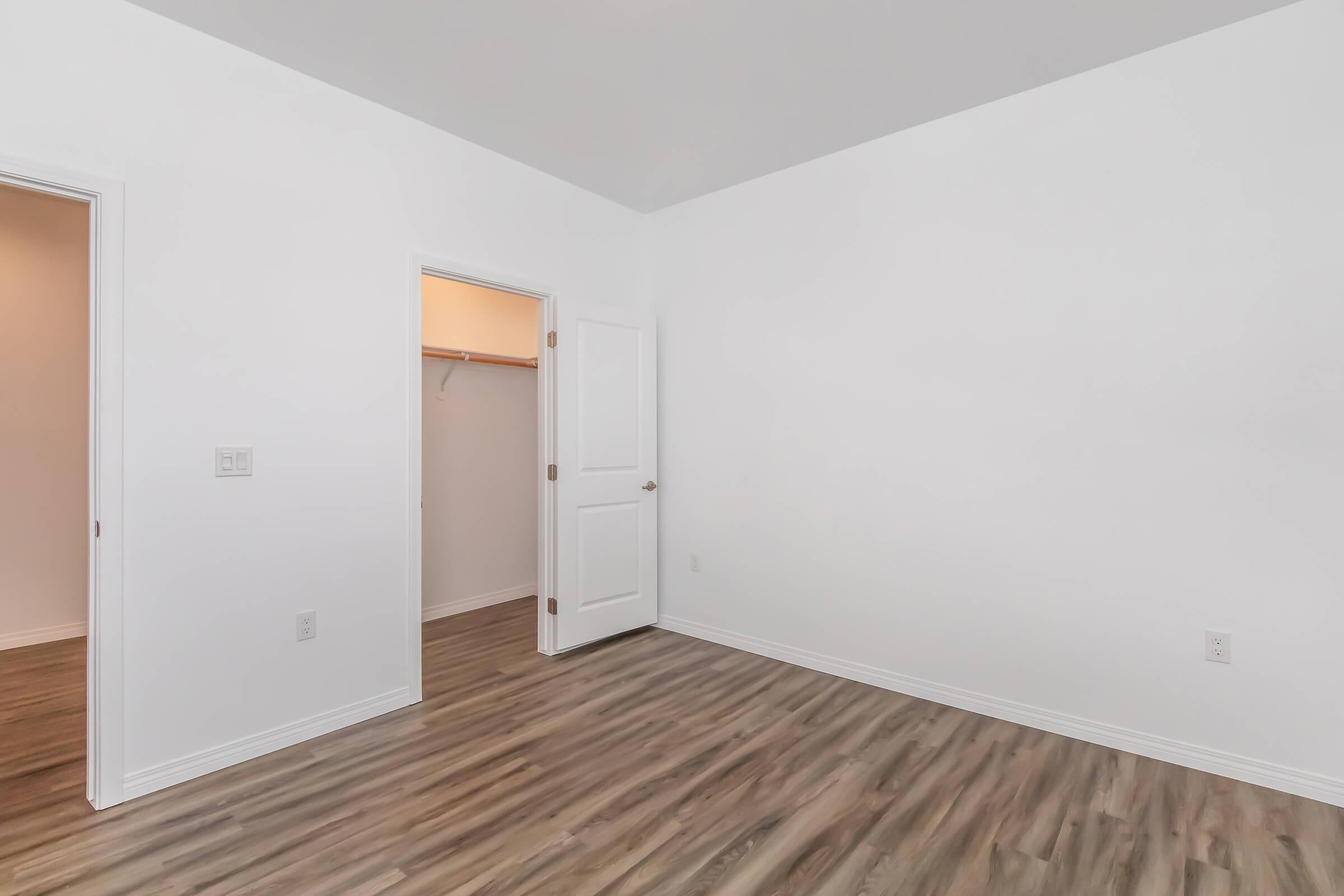A clean, empty room featuring light-colored walls and a wooden floor. There is a closed door on the right leading to a closet and another door on the left. The room is well-lit and has a minimalist appearance, making it suitable for various purposes.