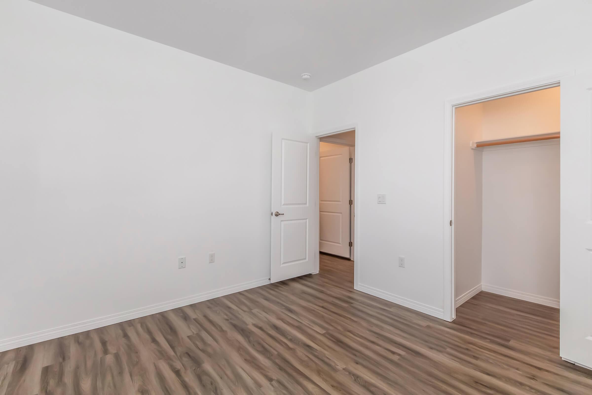 A bright, empty room with white walls and a light wood floor. One door leads to a closet, while another door opens to an adjoining space. The room has no furniture, allowing for various design possibilities.