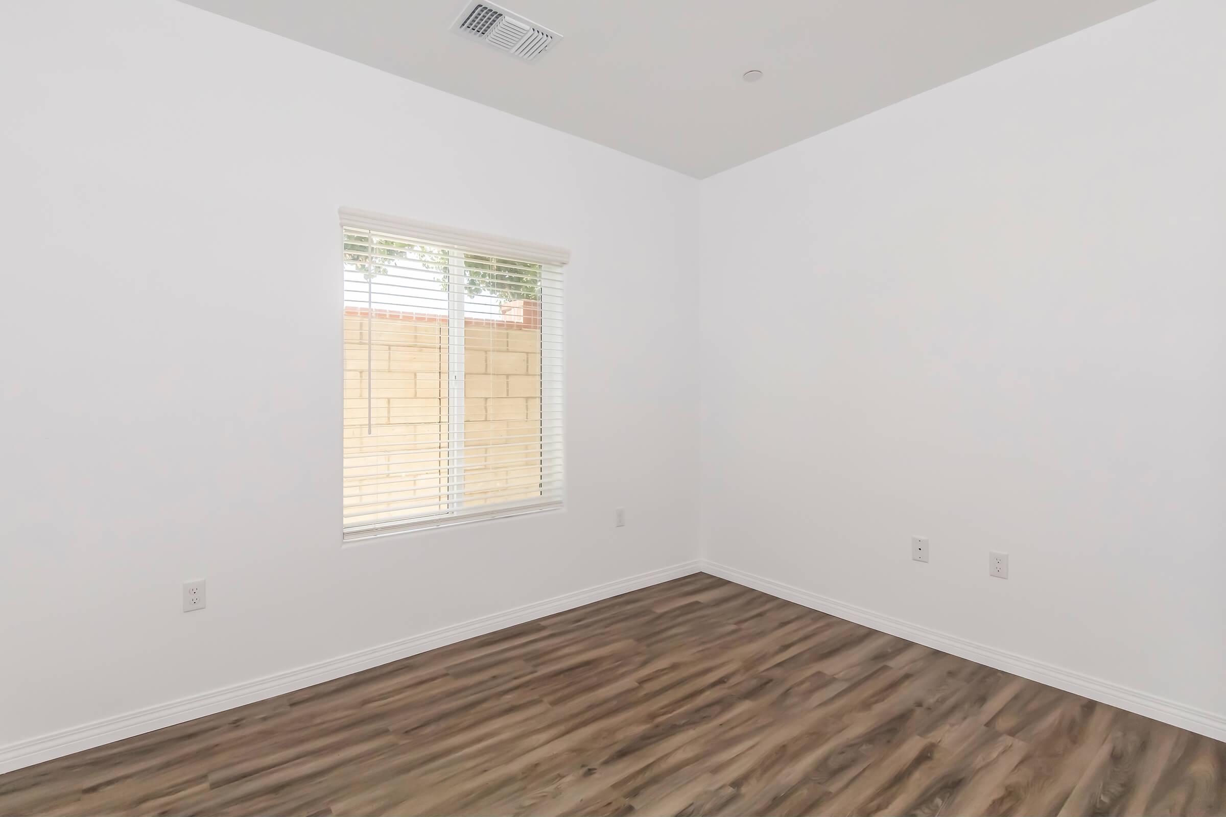 A minimalistic empty room with white walls and wooden flooring. There is a window with blinds on one wall, allowing natural light to enter. The room features no furniture or decorations, emphasizing a clean and open space.