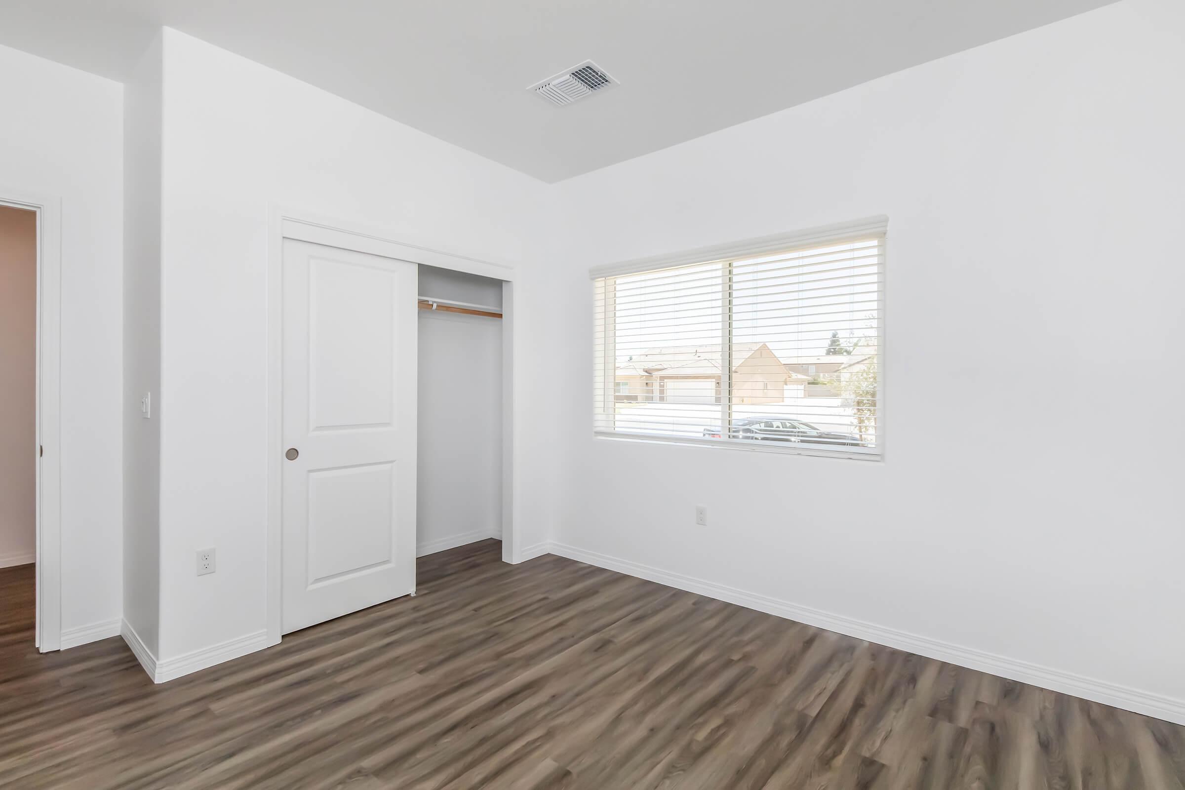 A bright, empty room with white walls and a window featuring blinds. The floor is made of wood-like laminate. There is a closet with sliding doors on one side and a doorway leading to another room. Natural light comes through the window, illuminating the space.