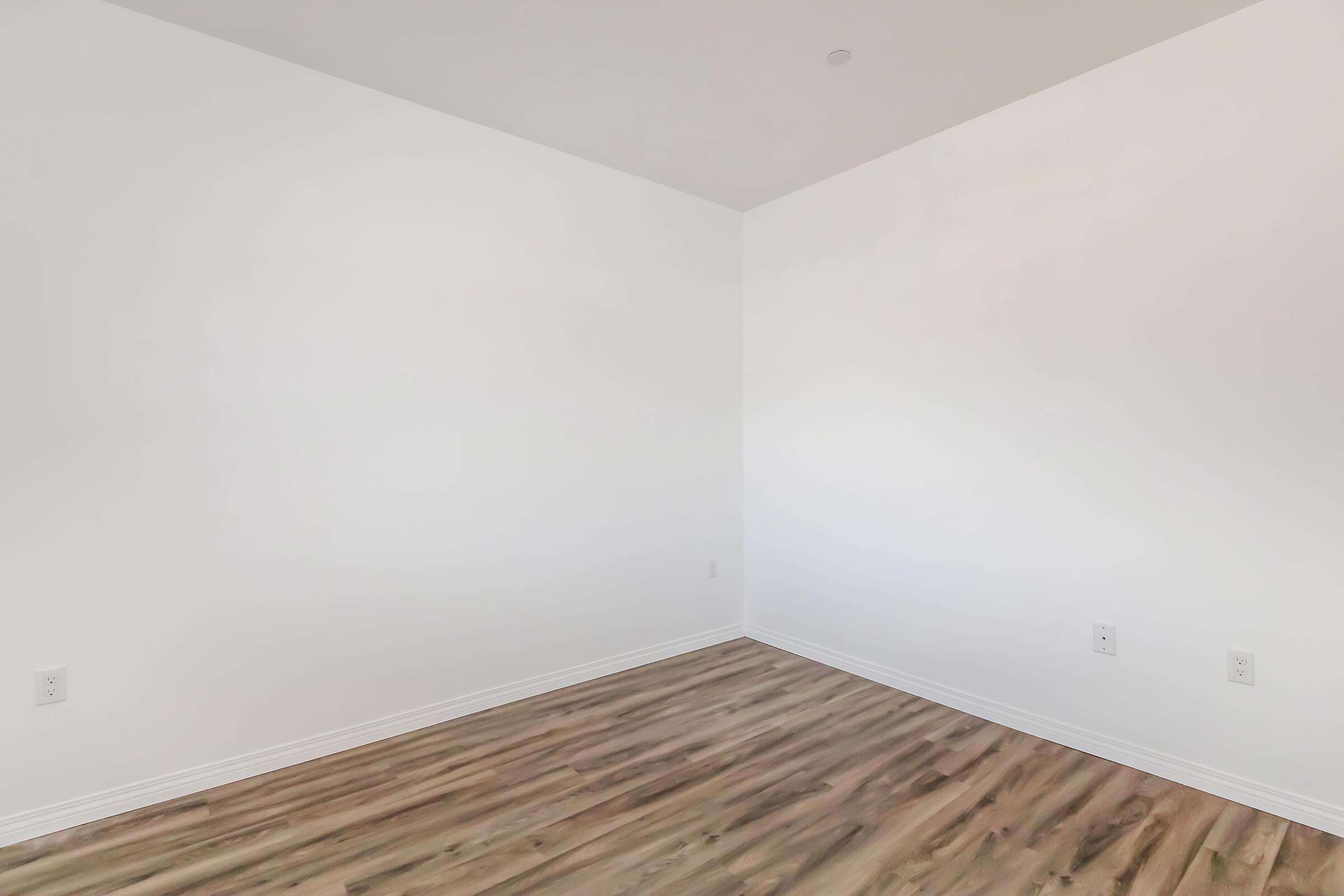 An empty room with white walls and a wooden floor. There are two electrical outlets visible on the wall, and the space is well-lit with natural light, creating a clean and minimalist atmosphere.