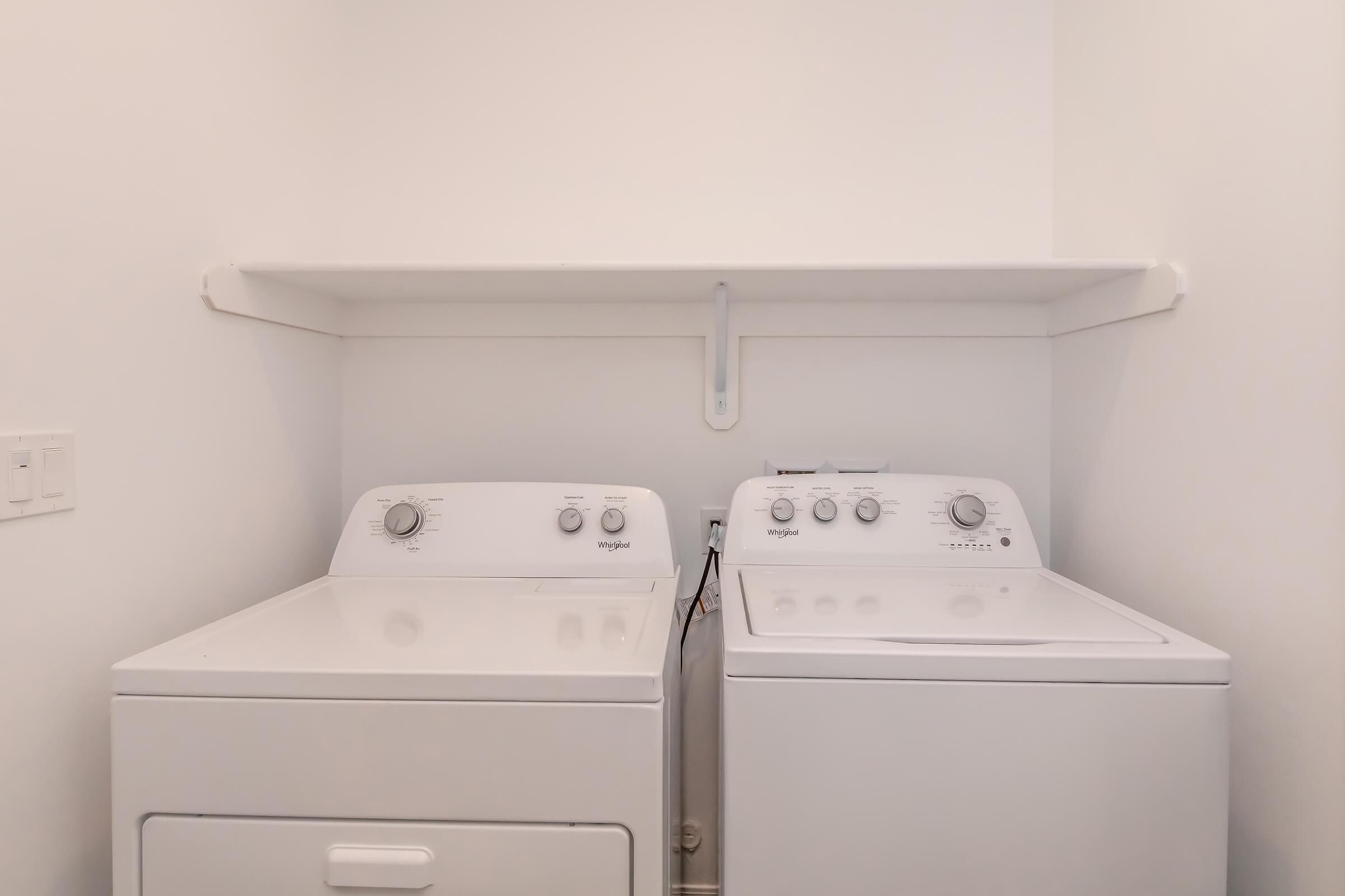 A clean, modern laundry room featuring a white washer and dryer side by side. The appliances have simple dials and controls. Above them is a minimalist shelf against a plain white wall, providing additional storage space. The floor is likely tiled or finished, enhancing the tidy appearance of the space.