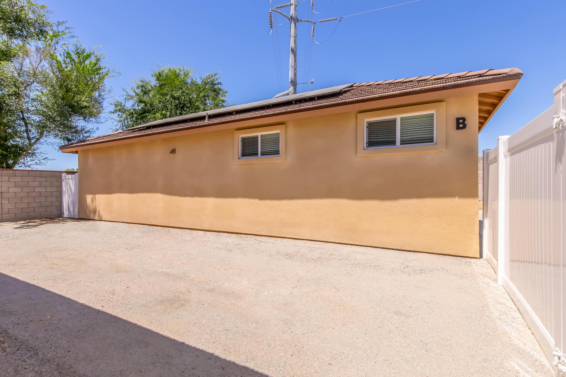 A quiet, empty side of a house with a beige exterior, featuring two windows on the wall and a surrounding gravel area. The image captures a clear blue sky and tall trees in the background, along with power lines visible above the roof. The property appears well-maintained and spacious.