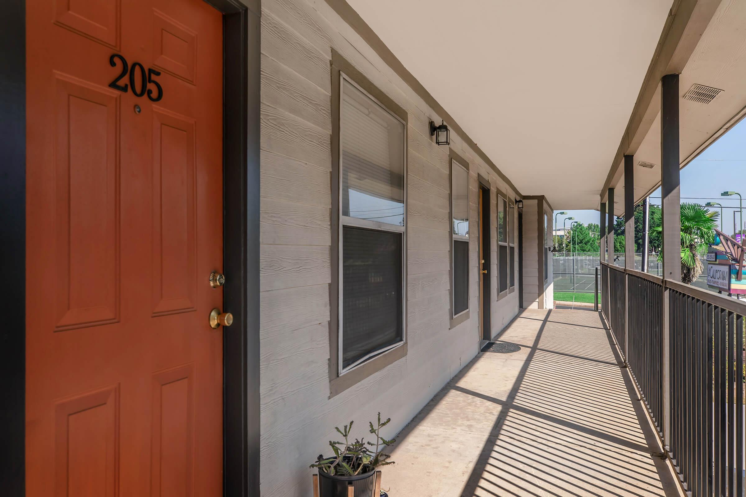 View of a second-floor apartment corridor featuring a bright orange door labeled "205." The walkway is lined with windows and a black railing, with sunlight casting shadows on the ground. A small potted plant is visible near the door, adding a touch of greenery to the entrance.