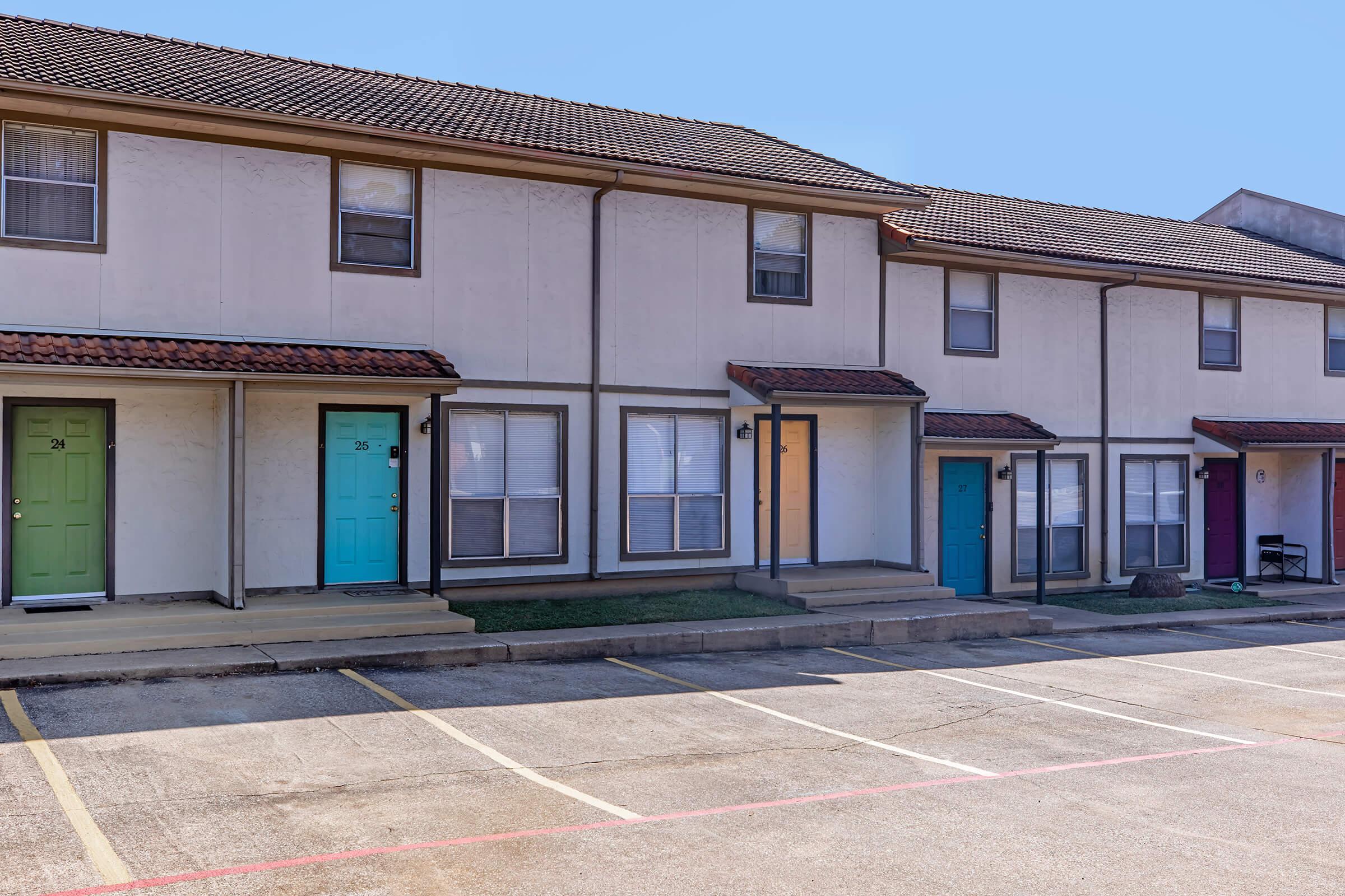 Row of two-story townhouses with numbered doors in various colors, including teal, yellow, and purple. The buildings have a simple, modern design with a concrete walkway leading to the entrances. An empty parking lot is visible in front, and the sky is clear and blue.