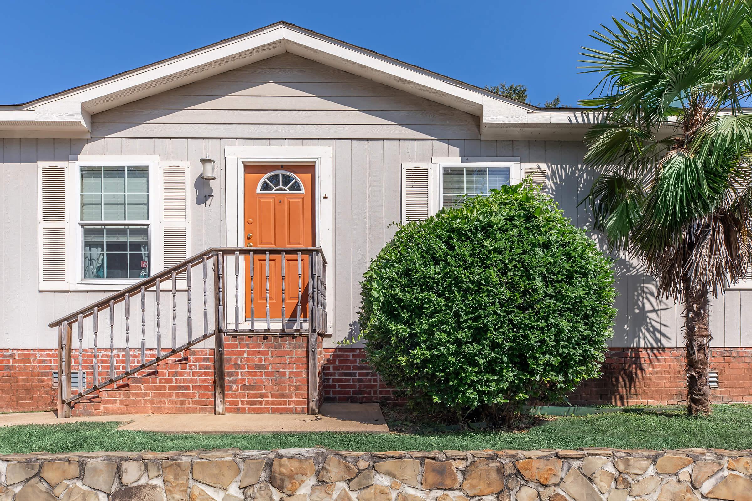 A single-story house with light gray siding and a brown front door. There is a small wooden staircase leading to the entrance. The front yard features a neatly trimmed bush and palm tree, with a stone wall in the foreground. Clear blue sky above creates a bright atmosphere.