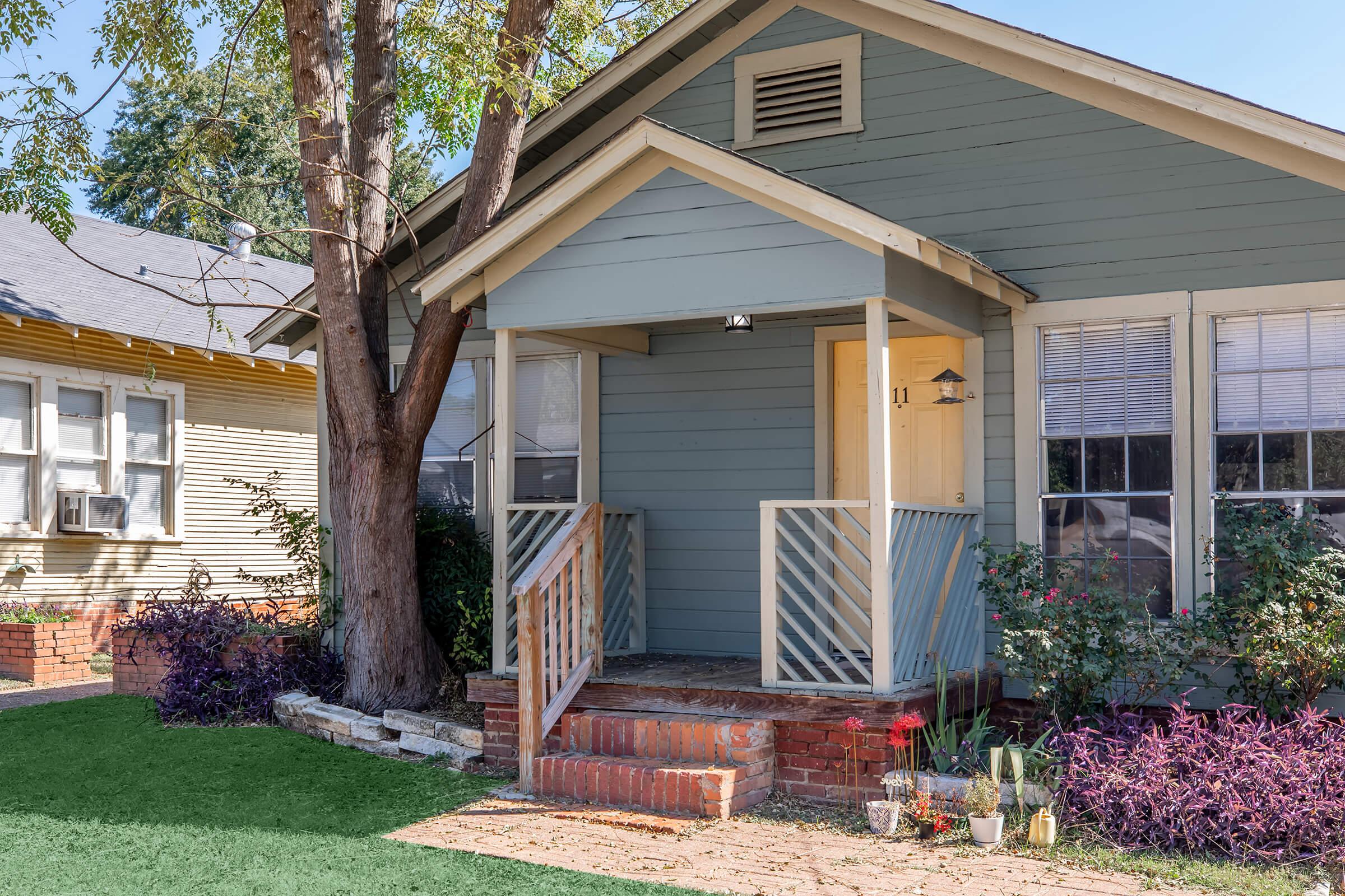 A charming, pale blue house with a front porch and white railing. The entrance features a cream-colored door. Surrounding the porch are green plants and flowers, with a large tree nearby. The yard has a neat, brick pathway leading up to the porch. Other houses can be seen in the background.