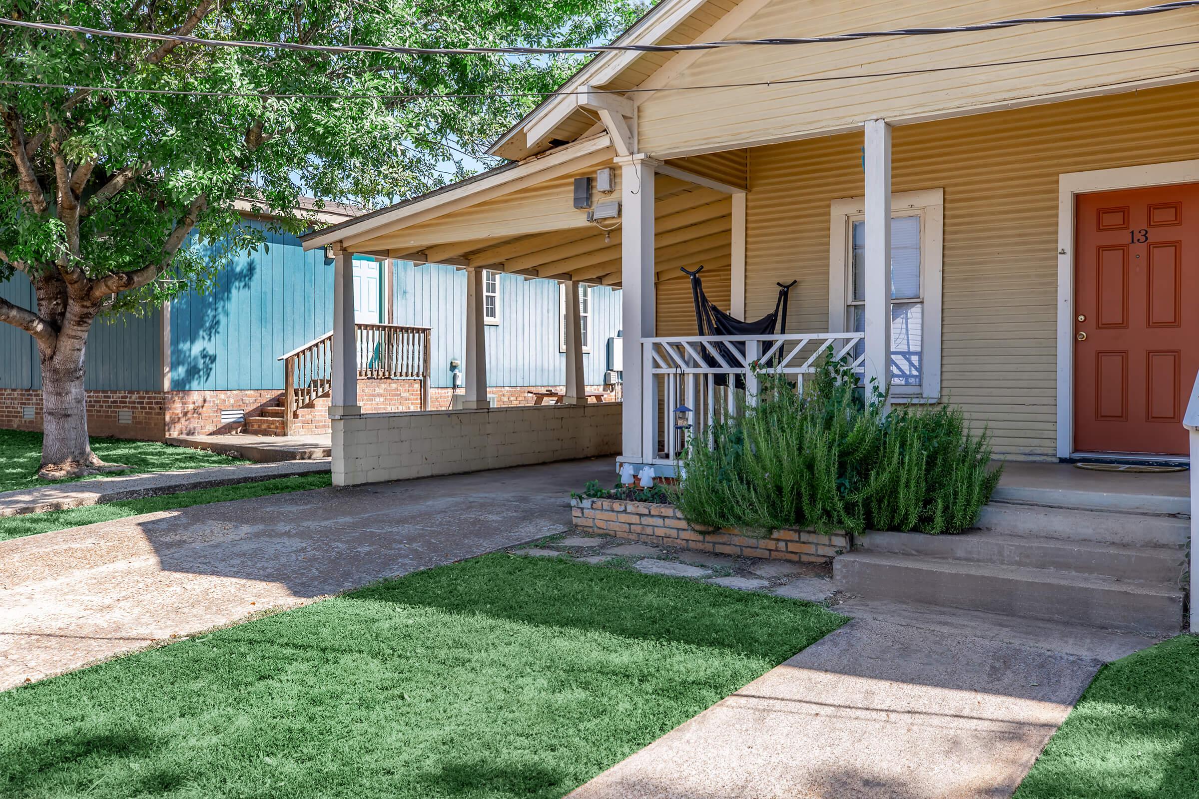 A residential exterior view showcasing a bungalow with a yellow facade and a welcoming front porch featuring a door numbered 13. The porch is decorated with plants, and the surrounding area includes grassy lawns and a tree. Neighboring houses are visible in the background, adding to the community atmosphere.