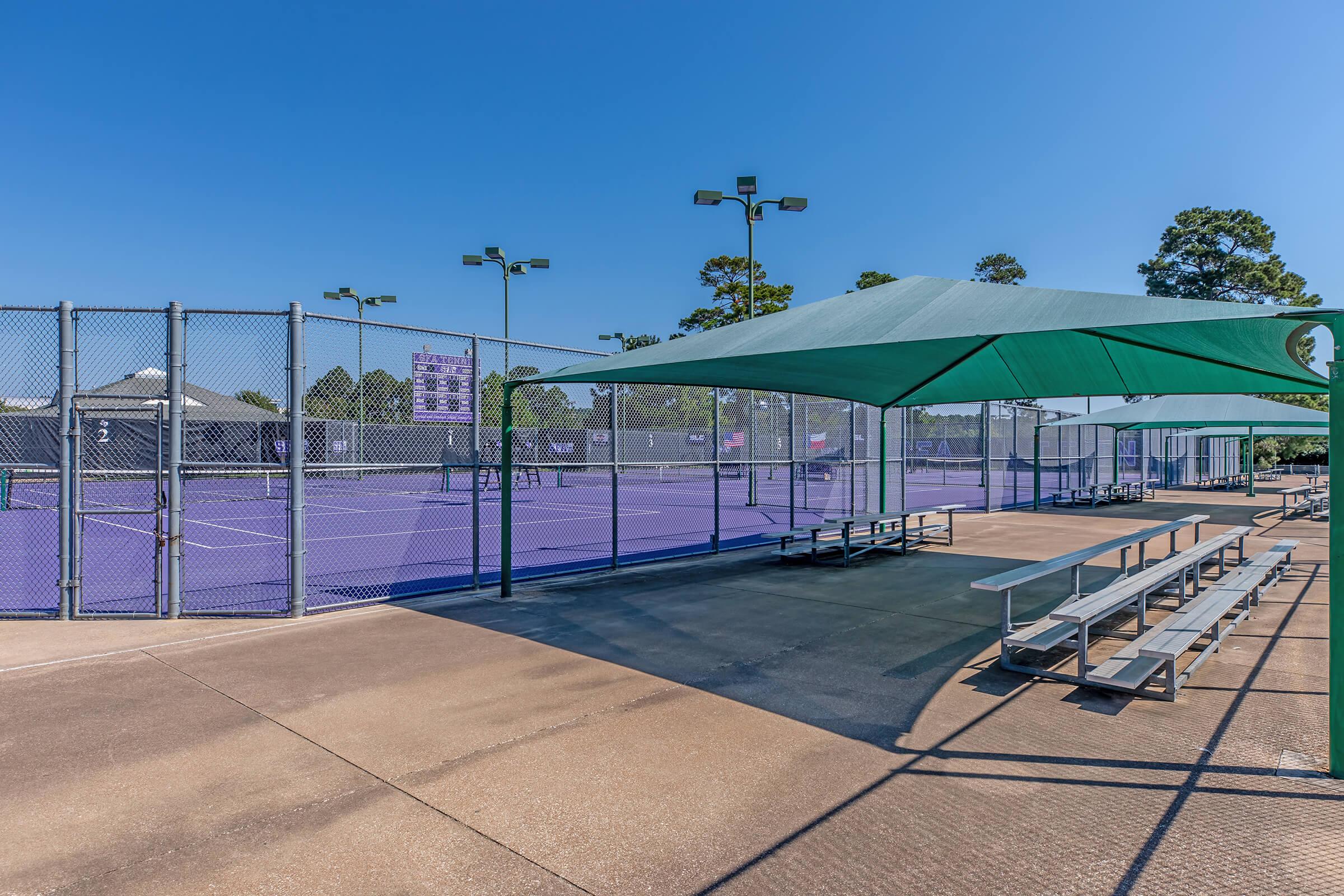 A well-lit outdoor tennis complex featuring multiple purple tennis courts. Shade structures provide cover for spectators, with benches visible along the side. The clear blue sky enhances the vibrant scene, making it ideal for recreational play and events.