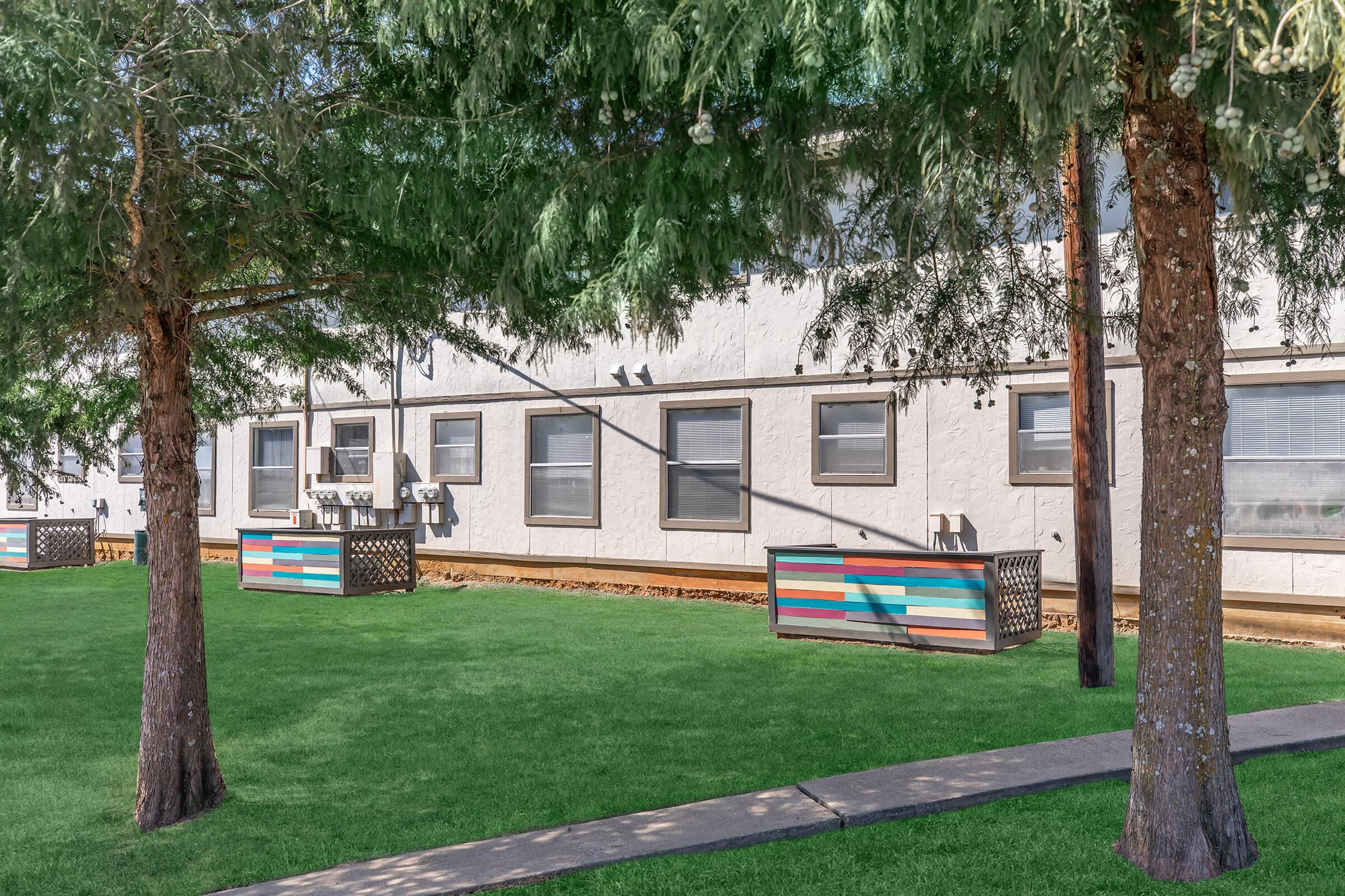 A lawn with green grass and colorful planter boxes in front of a white building. The area is shaded by tall trees, and there are windows visible on the building's exterior. The scene conveys a serene outdoor environment with a well-maintained landscape.