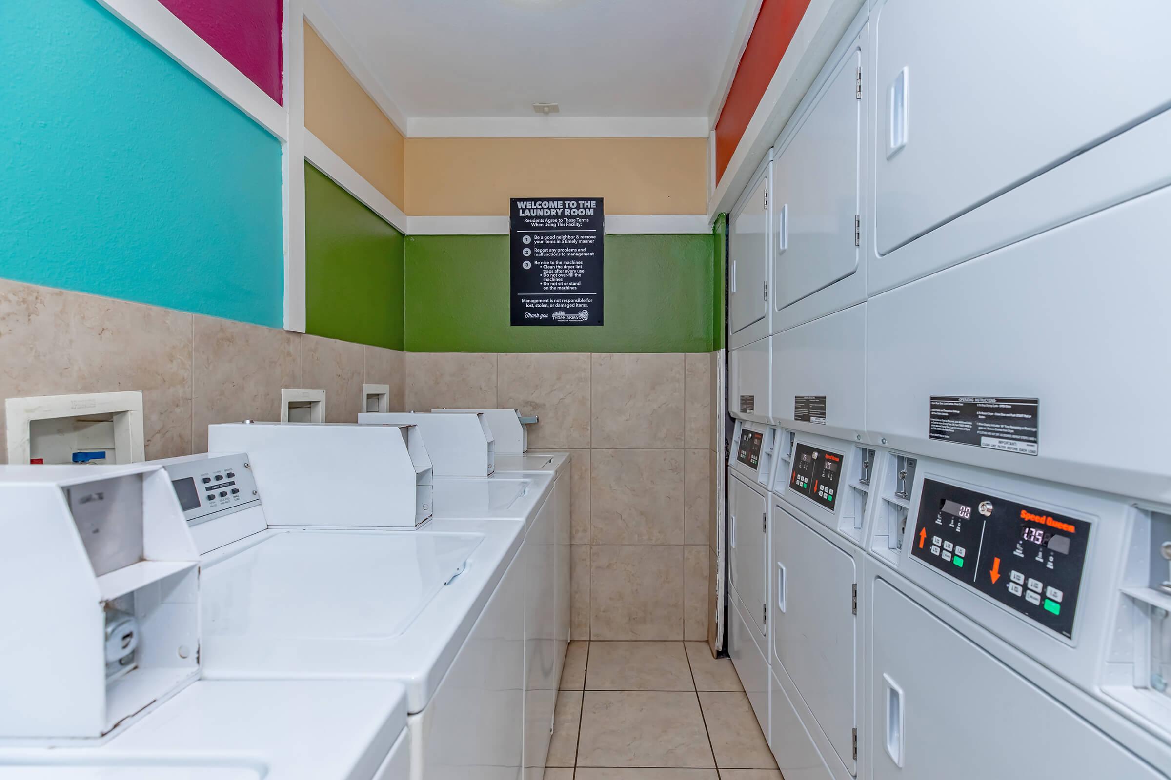 A well-lit laundry room featuring several stacked washing machines and dryers along one wall. Colorful walls in shades of teal, orange, and green add vibrancy. A sign on the wall provides instructions or information about the laundry facilities. The floor is tiled, creating a clean and organized space.