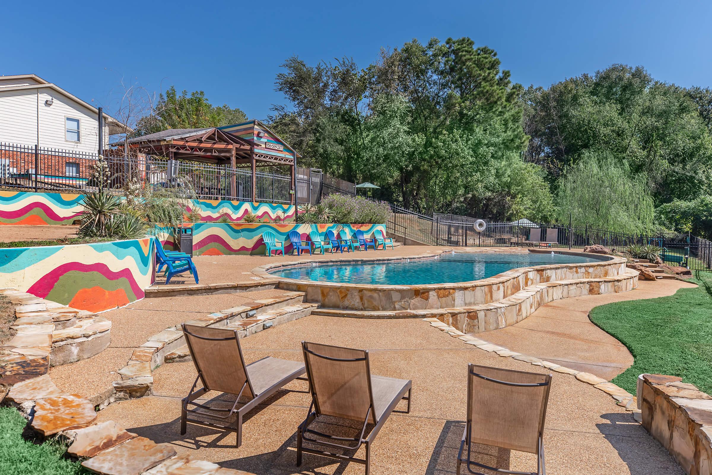 A vibrant outdoor swimming pool area featuring colorful painted walls, lounge chairs, and landscaped greenery. The pool is surrounded by a stone patio, with a nearby shaded structure and more seating. The scene is set against a clear blue sky and lush trees in the background.