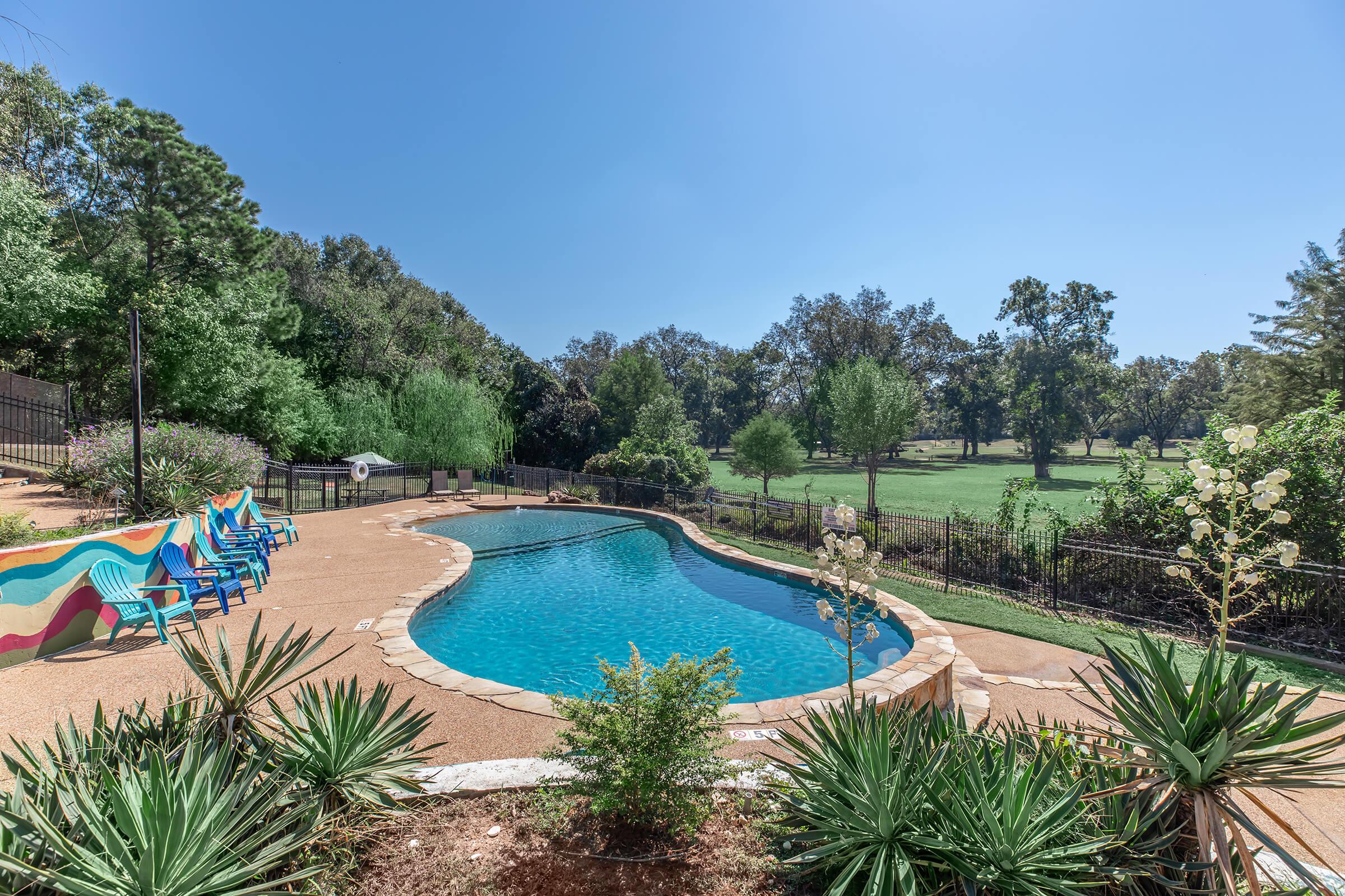 A serene outdoor swimming pool surrounded by lush greenery and landscaped gardens. Colorful lounge chairs line one side of the pool, and a fence marks the edge of the property, with rolling hills visible in the background under a clear blue sky.