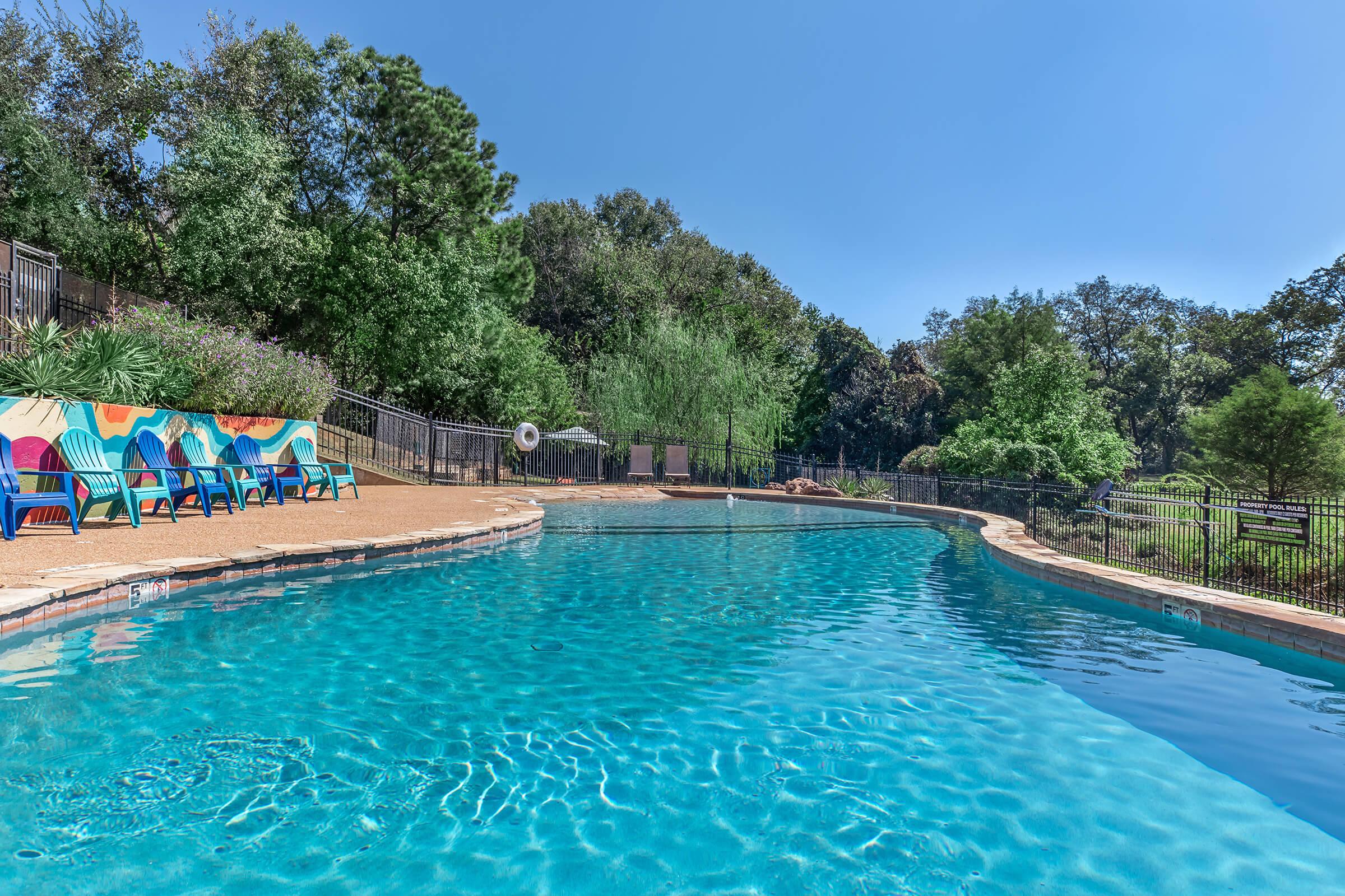 A clear, sparkling swimming pool surrounded by lush greenery and trees. Colorful lounge chairs line one side, and a black fence encloses the area. The sky is bright blue with no clouds, creating a warm and inviting atmosphere.