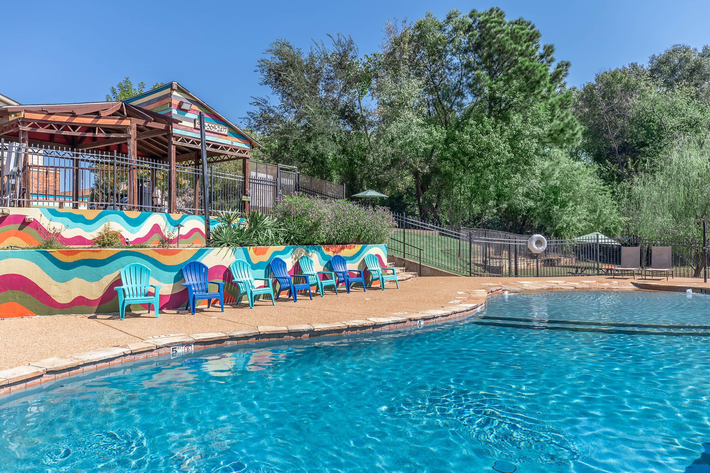 A vibrant outdoor pool area featuring a clear blue pool surrounded by colorful adirondack chairs. In the background, there's a shaded gazebo with artistic wave patterns on its side and lush greenery. The scene is bright and inviting, perfect for relaxation and leisure activities.