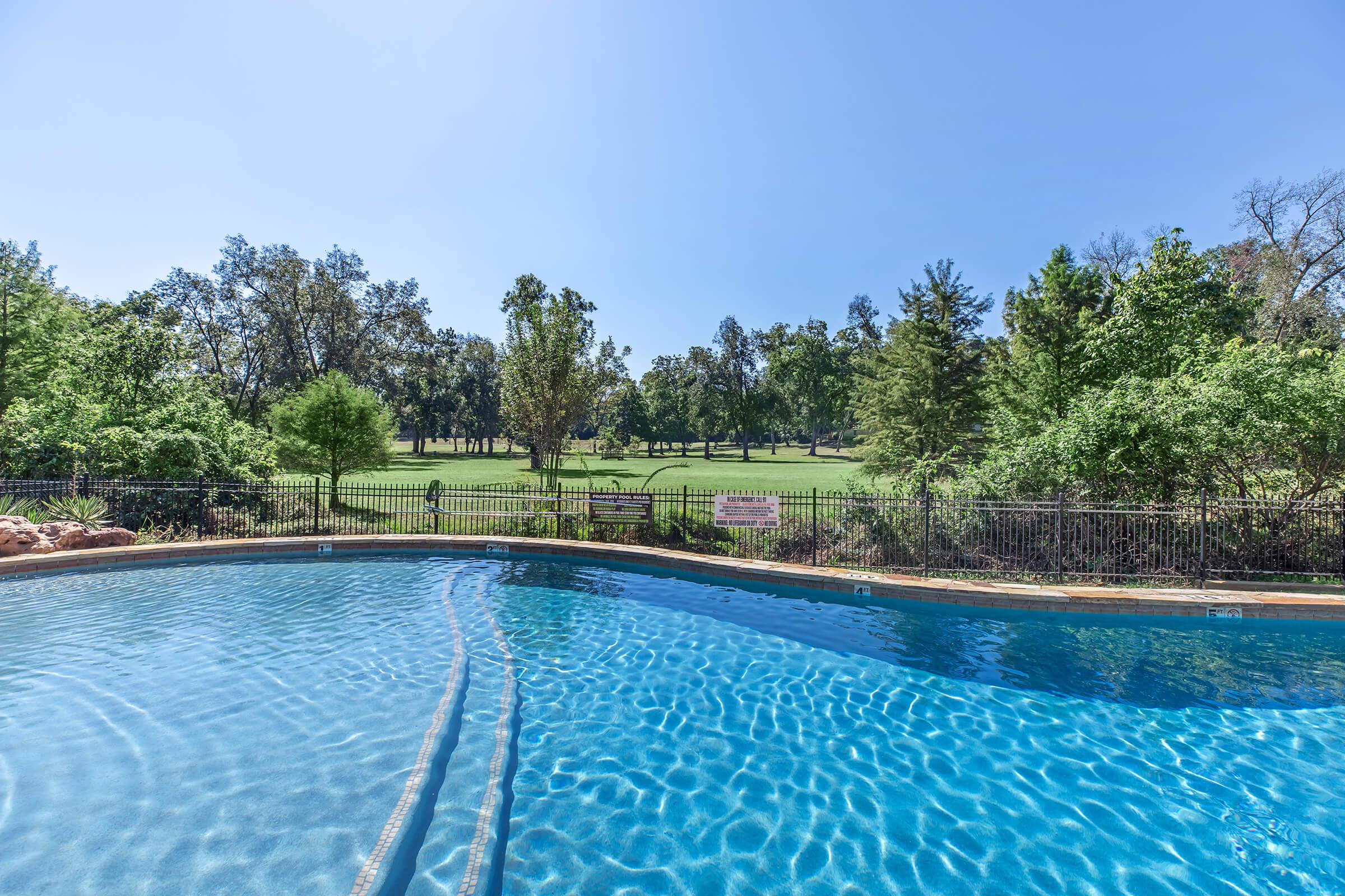 A tranquil swimming pool with clear blue water, surrounded by a black wrought-iron fence. Lush green trees and a spacious lawn are visible in the background under a clear blue sky. The scene conveys a serene outdoor setting ideal for relaxation and leisure.