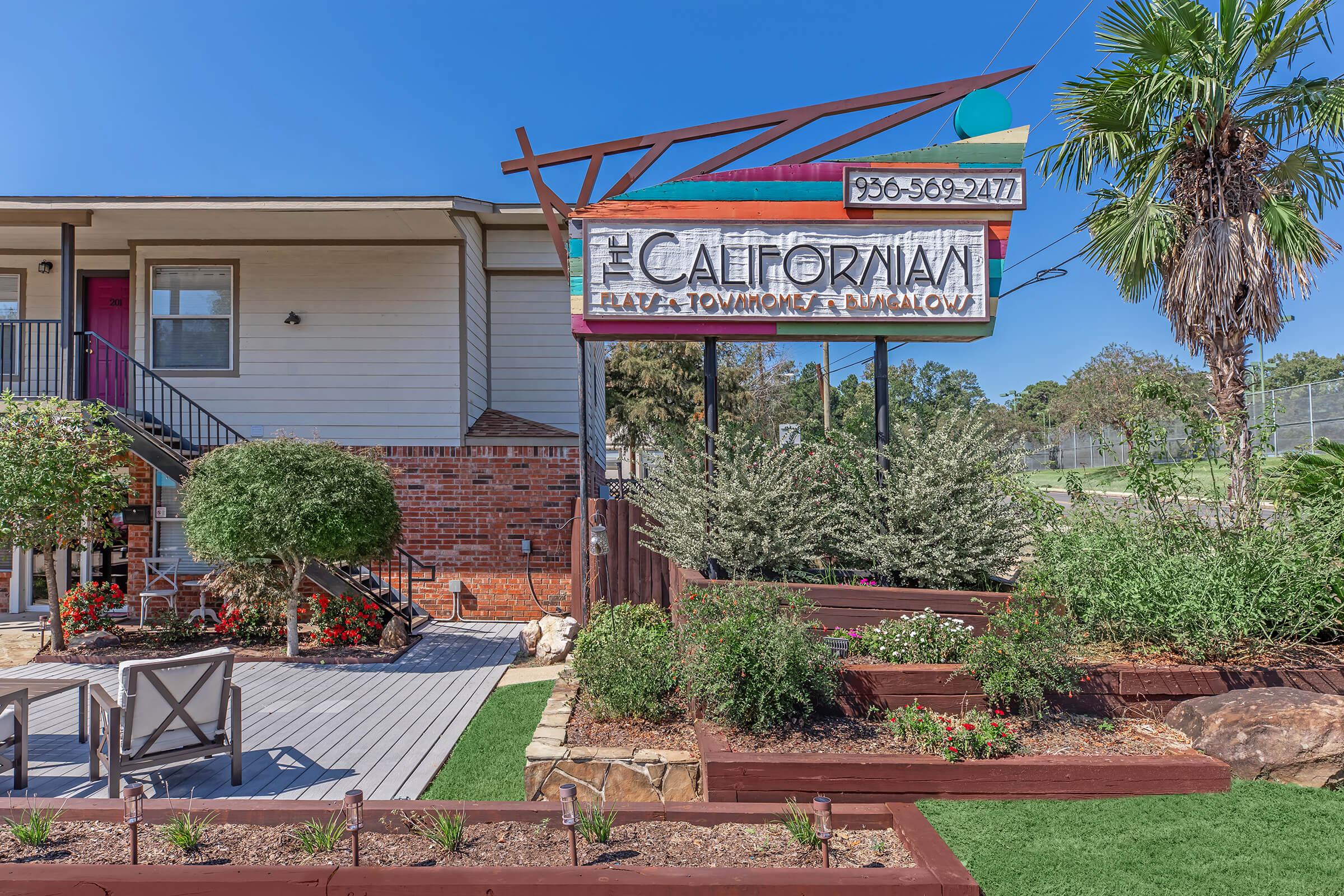 A colorful sign reading "The Californian" stands in front of a two-story building, surrounded by landscaped greenery and seating areas. The bright, blue sky and palm trees add to the vibrant setting, while the sign features contact information and a welcoming design.