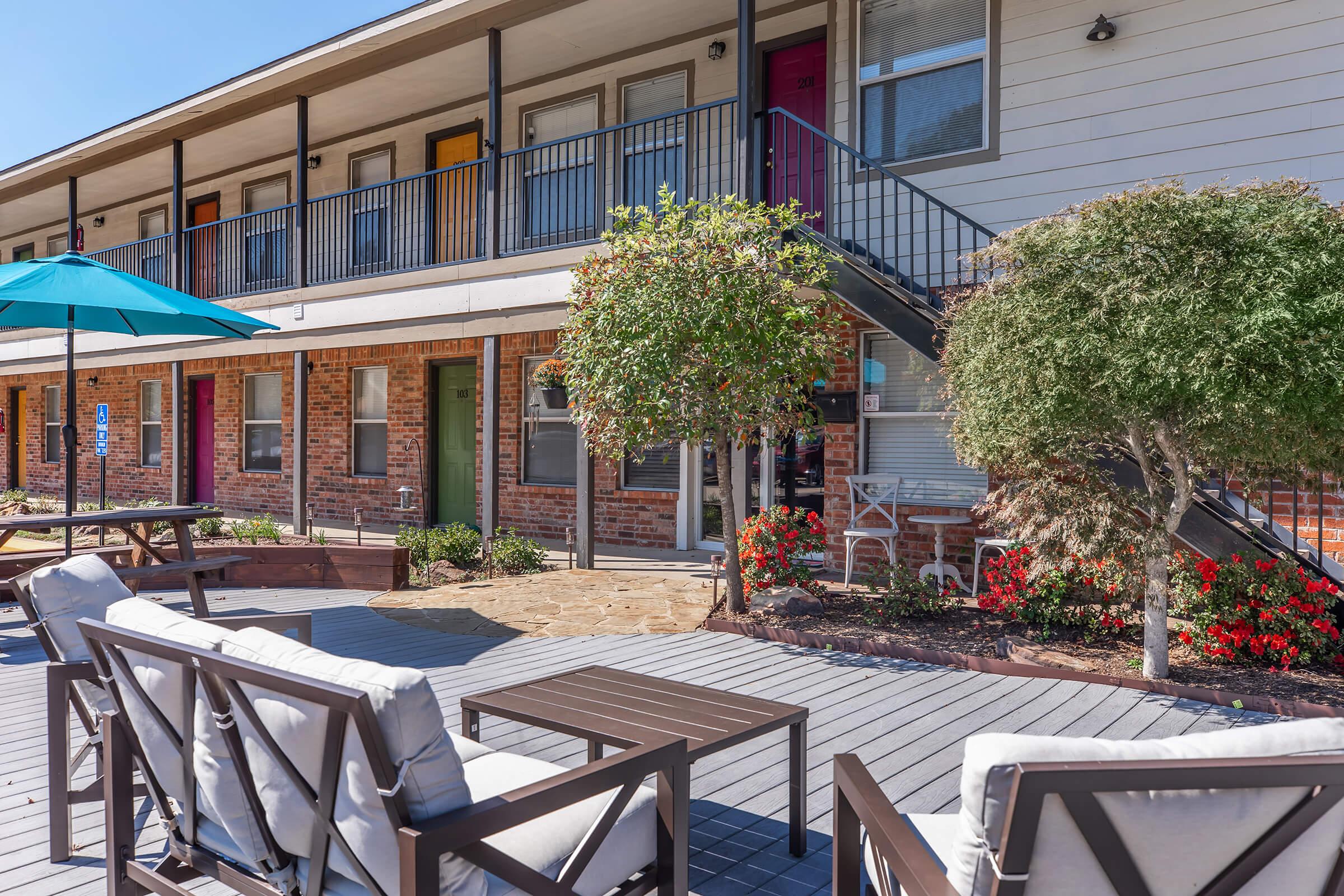 A cozy outdoor seating area with modern chairs and tables in front of a two-story building. The building features colorful doors in shades of green, red, and yellow. Lush shrubs and vibrant flowers enhance the inviting atmosphere, complemented by a large umbrella providing shade.