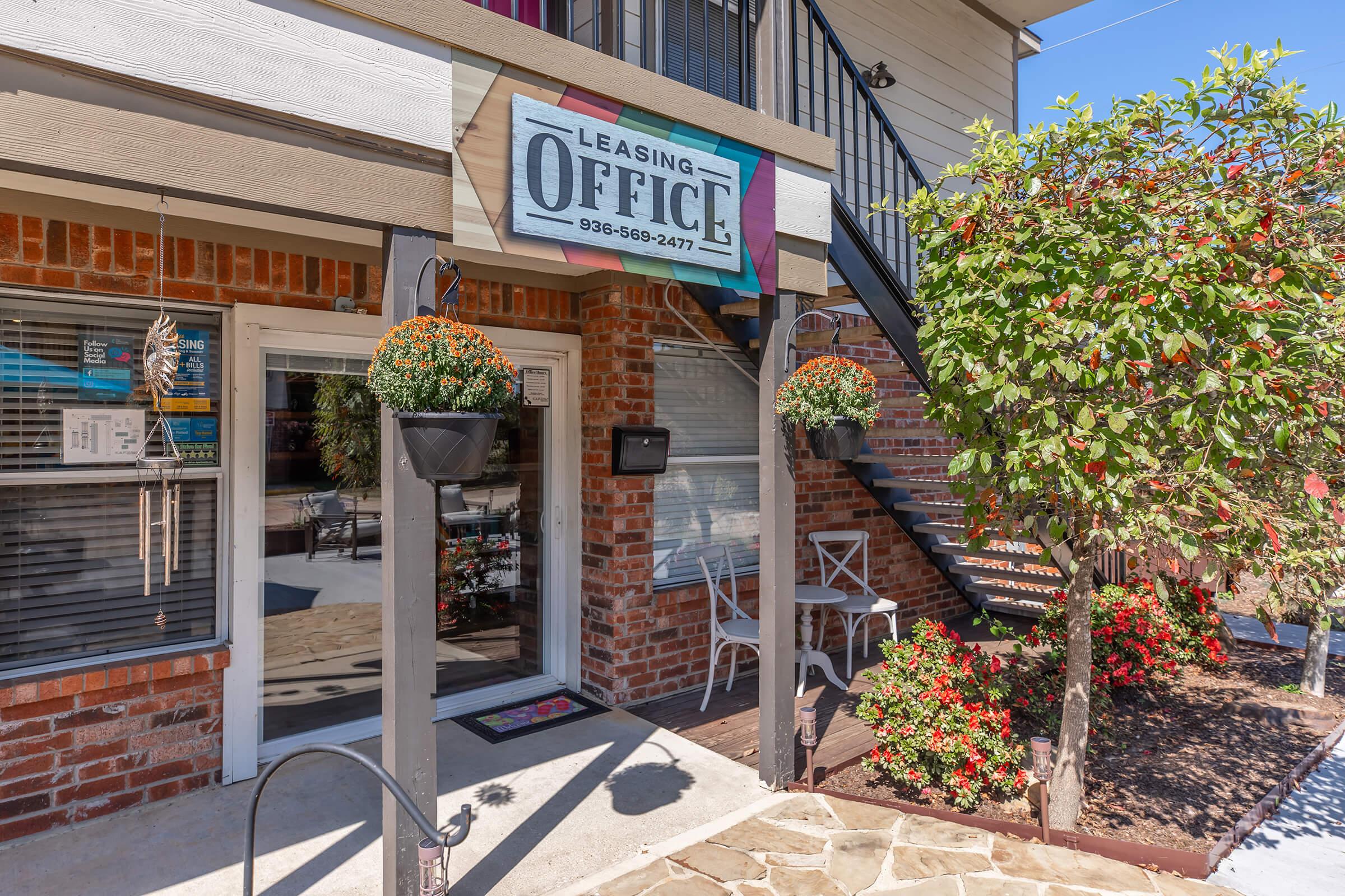 A leasing office entrance with a sign that reads "Leasing Office." The exterior features brick walls, a decorative staircase, hanging flower pots, and well-maintained landscaping with flowers and greenery. Sunlight brightens the scene, creating a welcoming atmosphere.