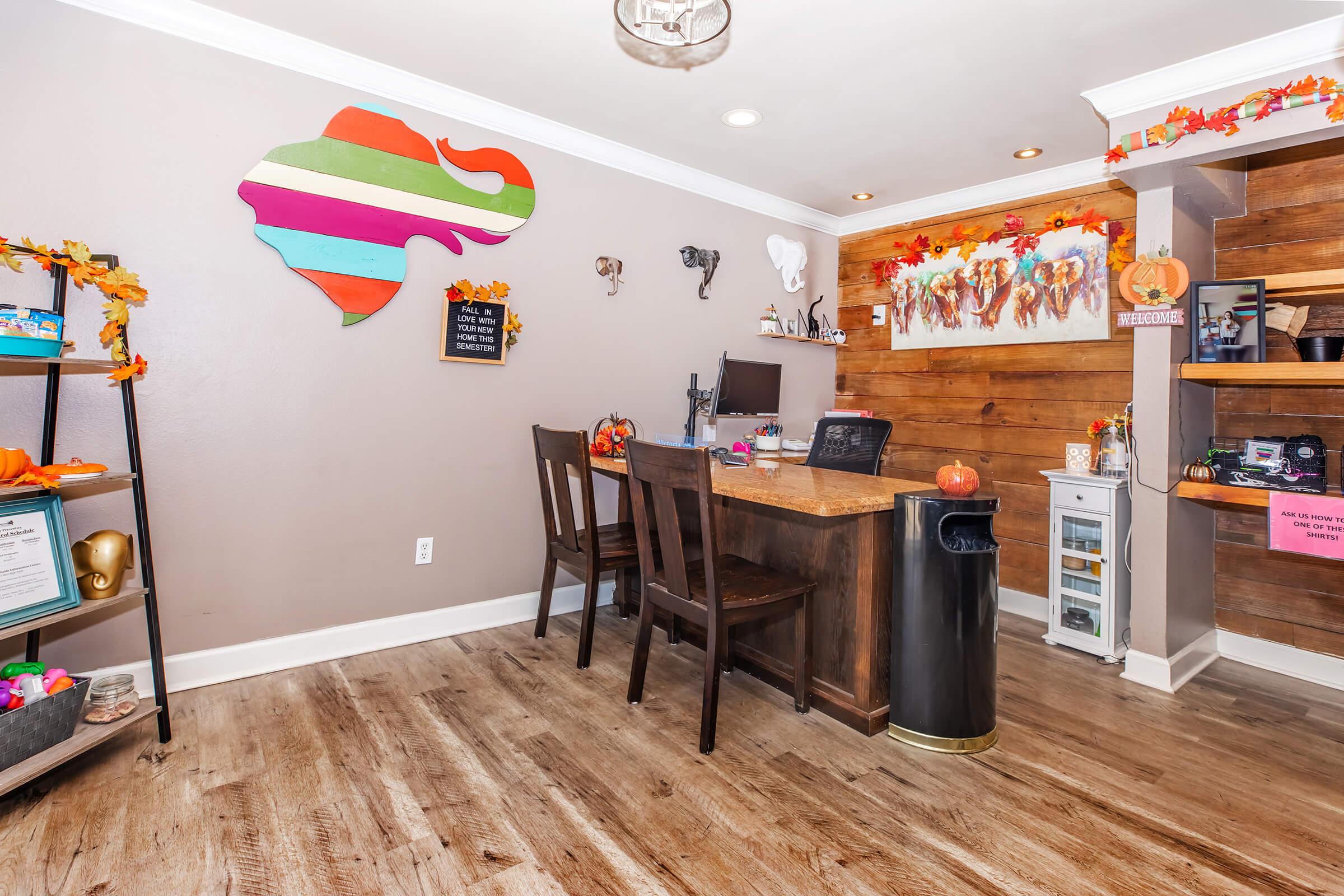 A cozy office space featuring a wooden desk with a computer, surrounded by autumn-themed decorations. A colorful wall art piece resembling a state outline is mounted on the wall. There are shelves with various items and a small trash can in the corner. The flooring is wooden, adding warmth to the environment.