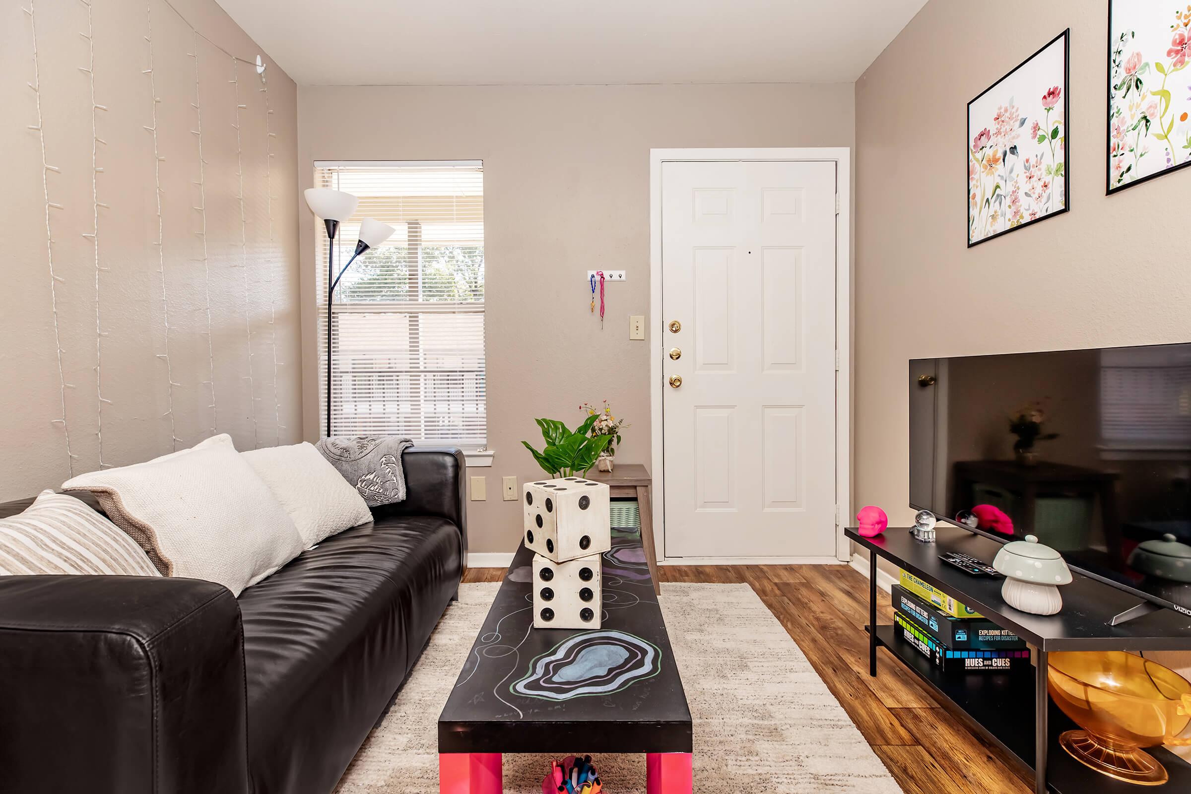 Cozy living room featuring a black sofa, a decorative coffee table with a colorful design, a TV stand with books and decor, and a door leading outside. Natural light streams in through a window adorned with light curtains. The walls are painted beige, with framed floral artwork accentuating the space.