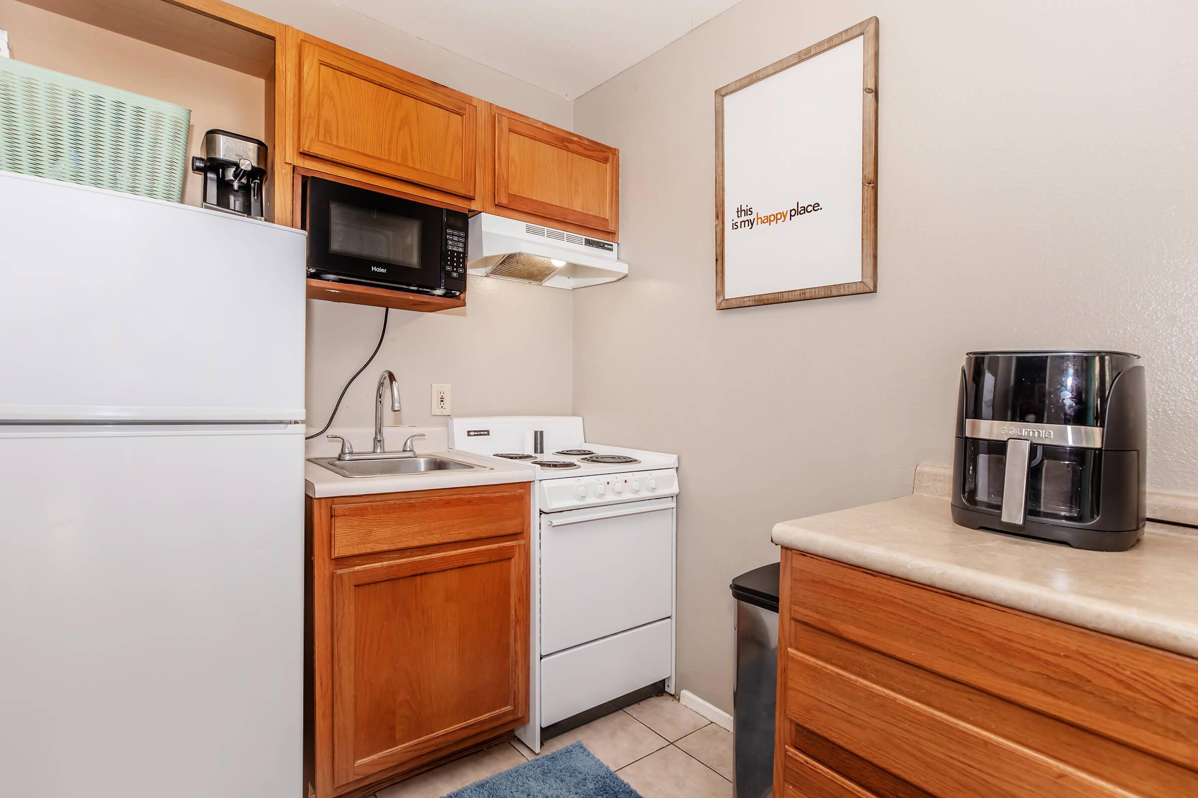 A compact kitchen featuring wooden cabinets, a white refrigerator, a microwave above the stove, a sink, and an electric cooktop. There is a countertop with a coffee maker and a framed sign on the wall that reads "It's a happy place." The floor is tiled, and the overall color scheme is light and airy.