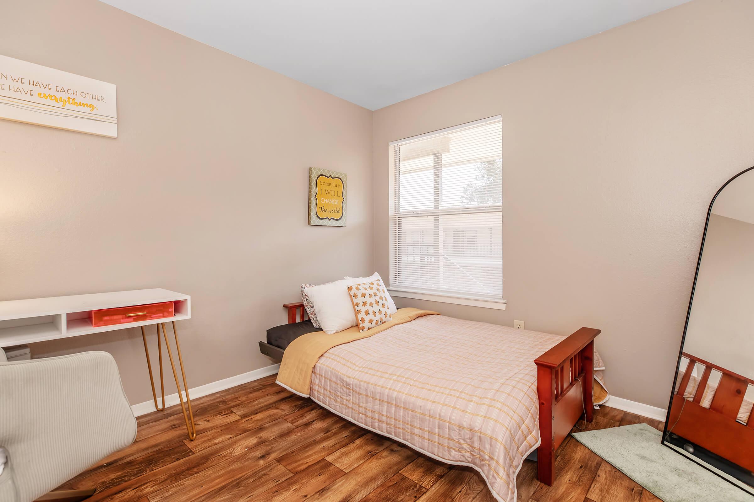 A cozy bedroom featuring a single bed with a beige blanket and decorative pillows, a small white desk with a chair, a full-length mirror, and a window allowing natural light. The walls are painted a light beige, and there’s a decorative sign on the wall. The floor is made of wooden planks.