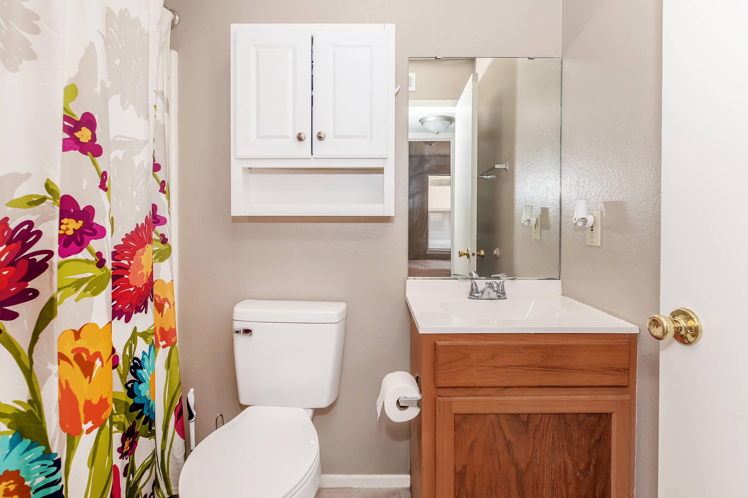 A small bathroom featuring a floral shower curtain, a white toilet, and a brown vanity with a sink. A cabinet above the vanity offers storage, and a mirror reflects the room's light. The walls are painted in a neutral tone, enhancing the airy feel of the space.