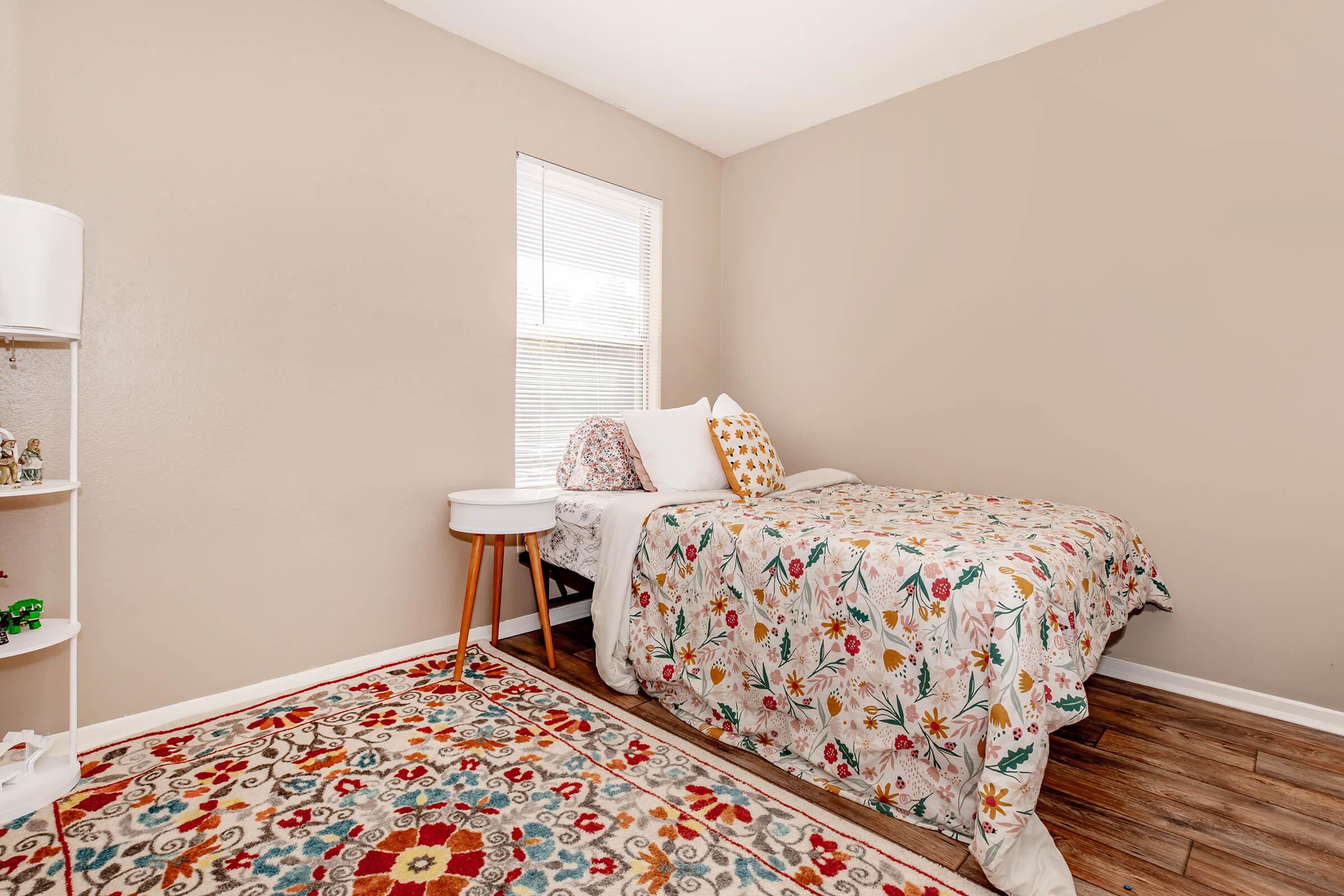 A cozy bedroom featuring a bed with a colorful floral patterned quilt, adorned with decorative pillows. Nearby is a small round bedside table and a bookshelf. The room has light beige walls and a window that allows natural light, with a vibrant patterned rug on the floor adding warmth to the space.