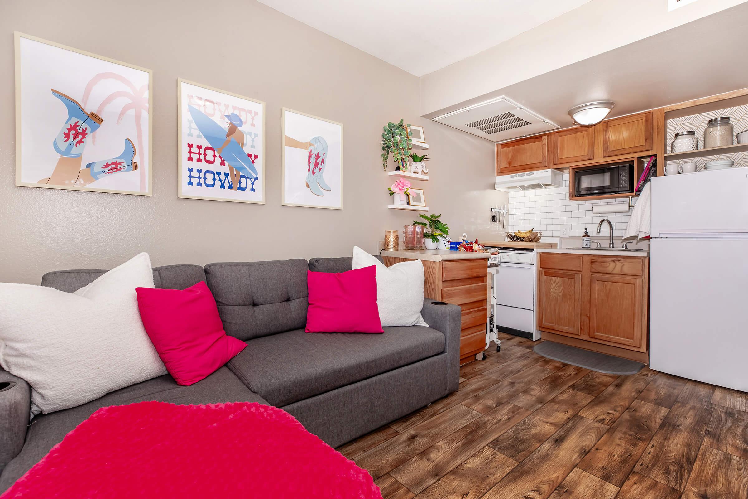 Cozy living space featuring a gray sofa with pink pillows, a wooden side table, and a kitchenette. The kitchen includes a white refrigerator, stove, and sink, surrounded by light-colored cabinetry. Three framed artwork pieces adorn the wall, adding a splash of color to the warm-toned room.