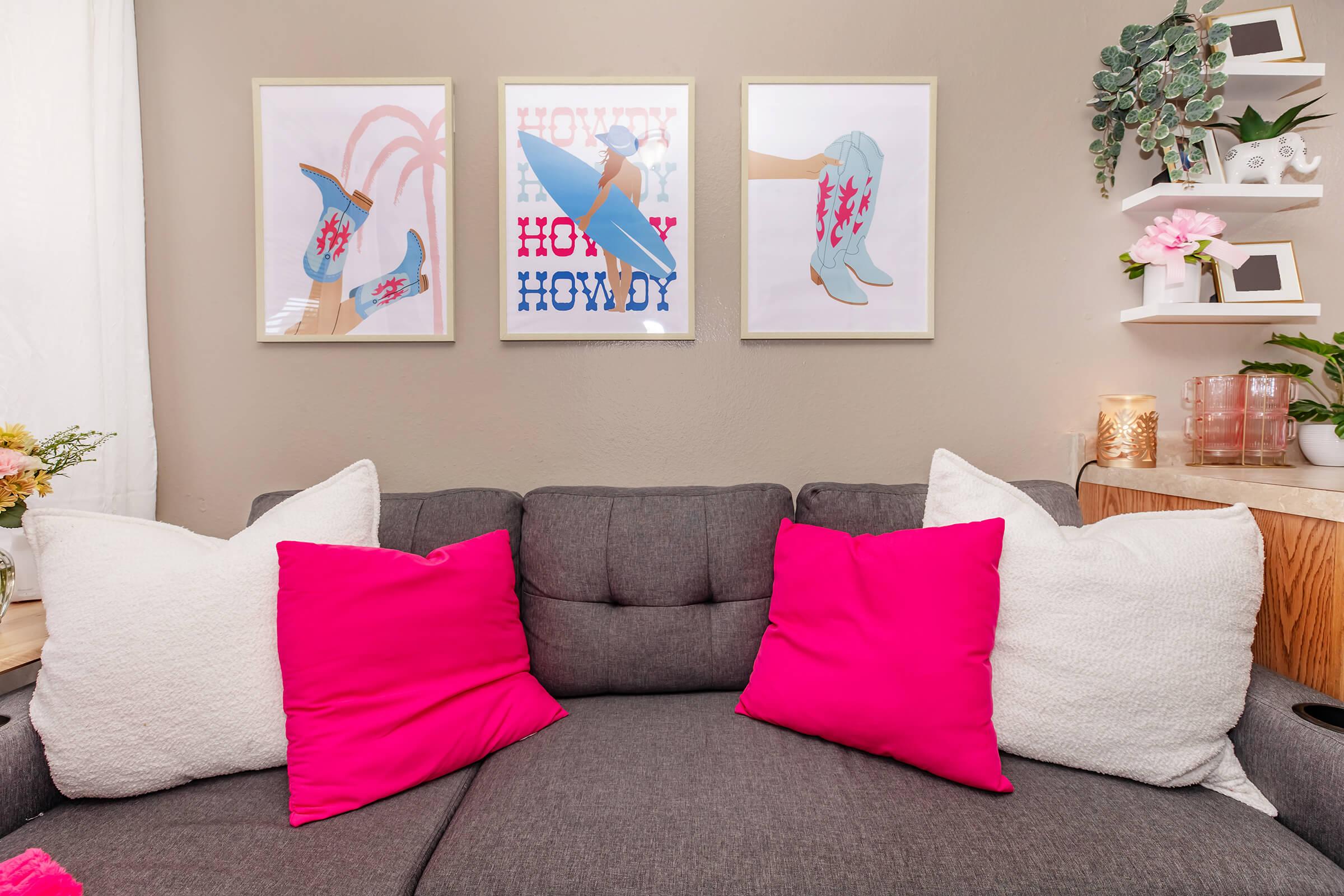 A cozy living room scene featuring a gray couch adorned with vibrant pink and white pillows. Above the couch are three framed art prints with a playful western theme, displaying images of cowboy boots and hats, adorned with the text "Howdy." The room has a warm and inviting atmosphere, enhanced by decorative plants and soft lighting.