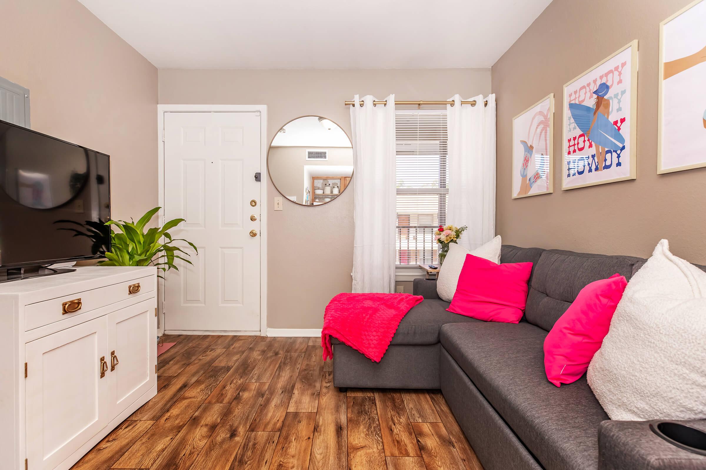 Cozy living room featuring a gray sectional sofa with bright pink cushions, a decorative pink throw blanket, and a white cabinet beneath a flat-screen TV. The space includes a round mirror, light curtains, and framed artwork on the walls, creating a welcoming and modern atmosphere.