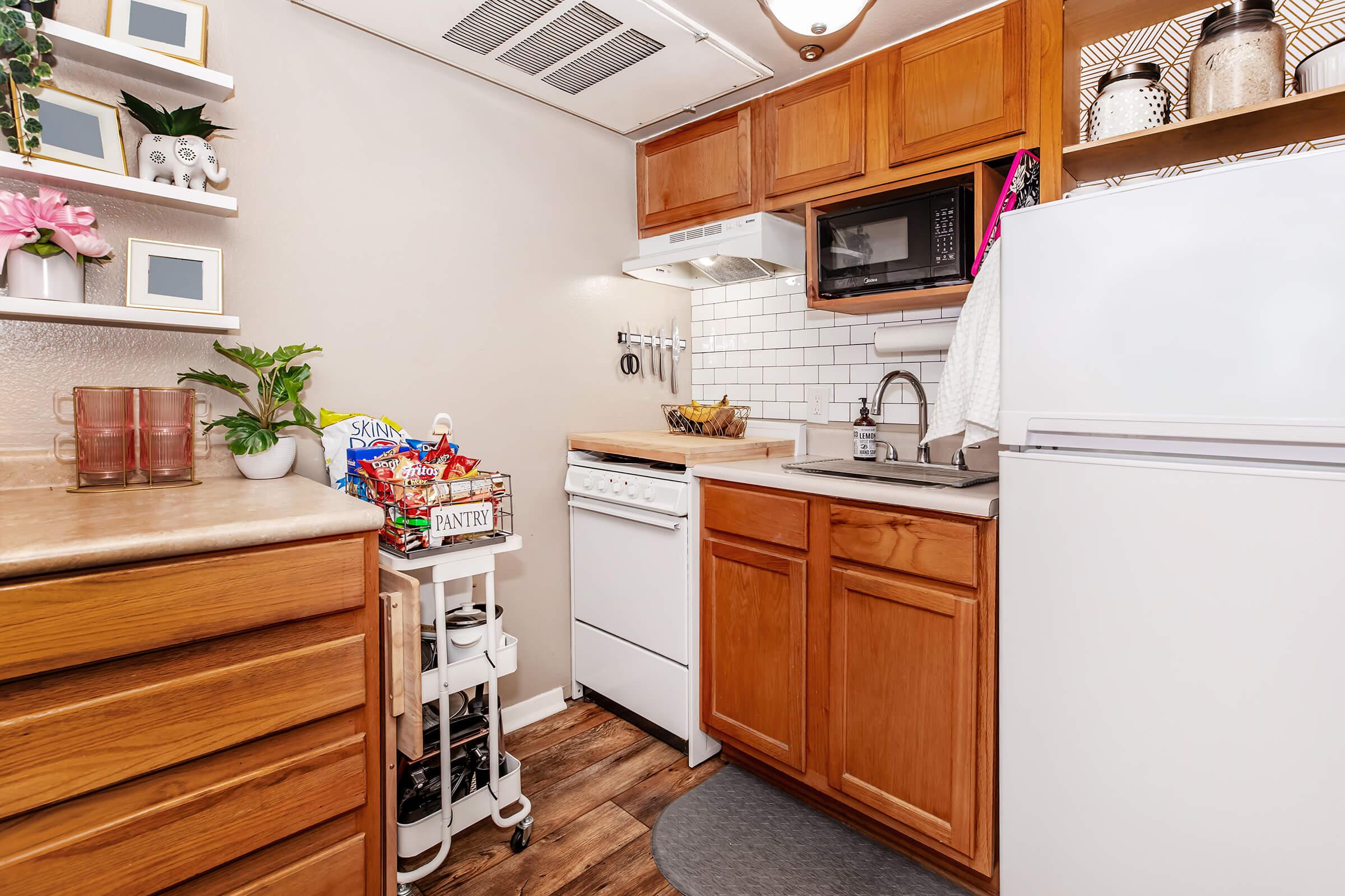 A compact kitchen featuring wooden cabinets, a white refrigerator, and a built-in microwave. The countertop holds a sink and a drying rack, with a small cart nearby filled with snacks. Decor elements include potted plants and framed pictures on the wall, creating a cozy atmosphere.