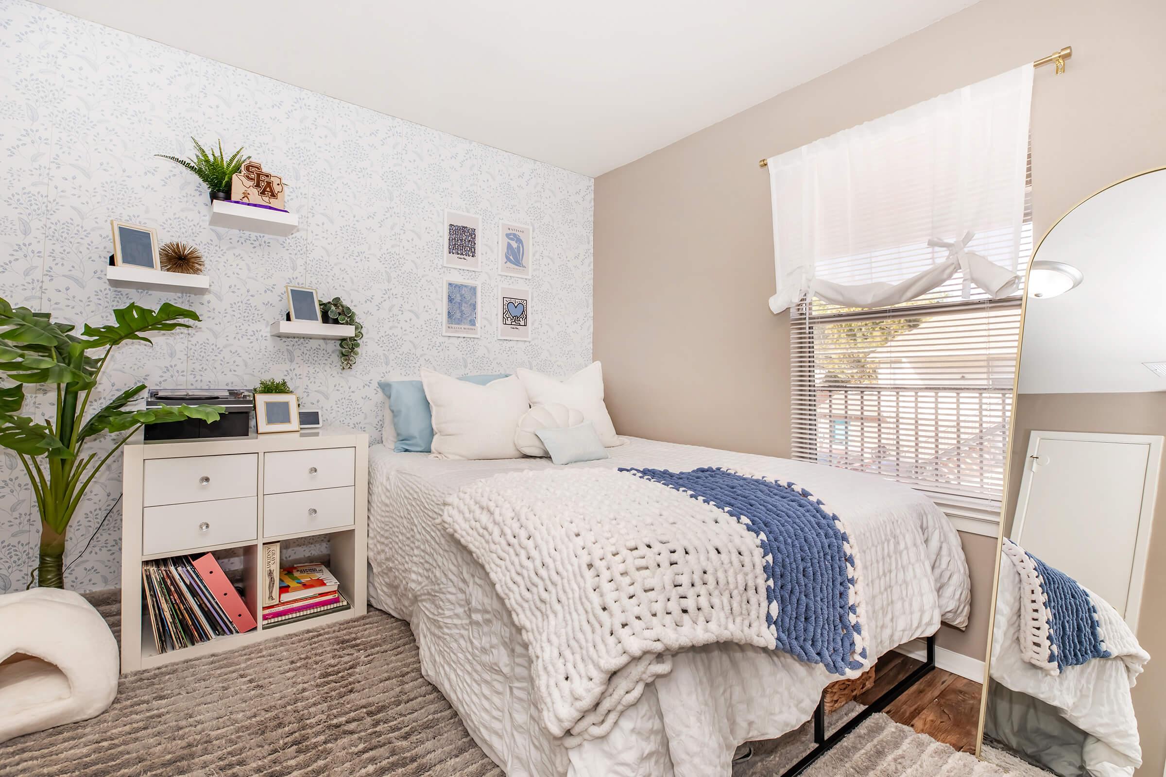 A cozy bedroom featuring a bed with a light-colored knitted blanket, decorative pillows, and a stylish wall with floral wallpaper. A small bookshelf displays books and plants, while a mirror reflects natural light from a nearby window with sheer curtains. A soft rug adds warmth to the space.