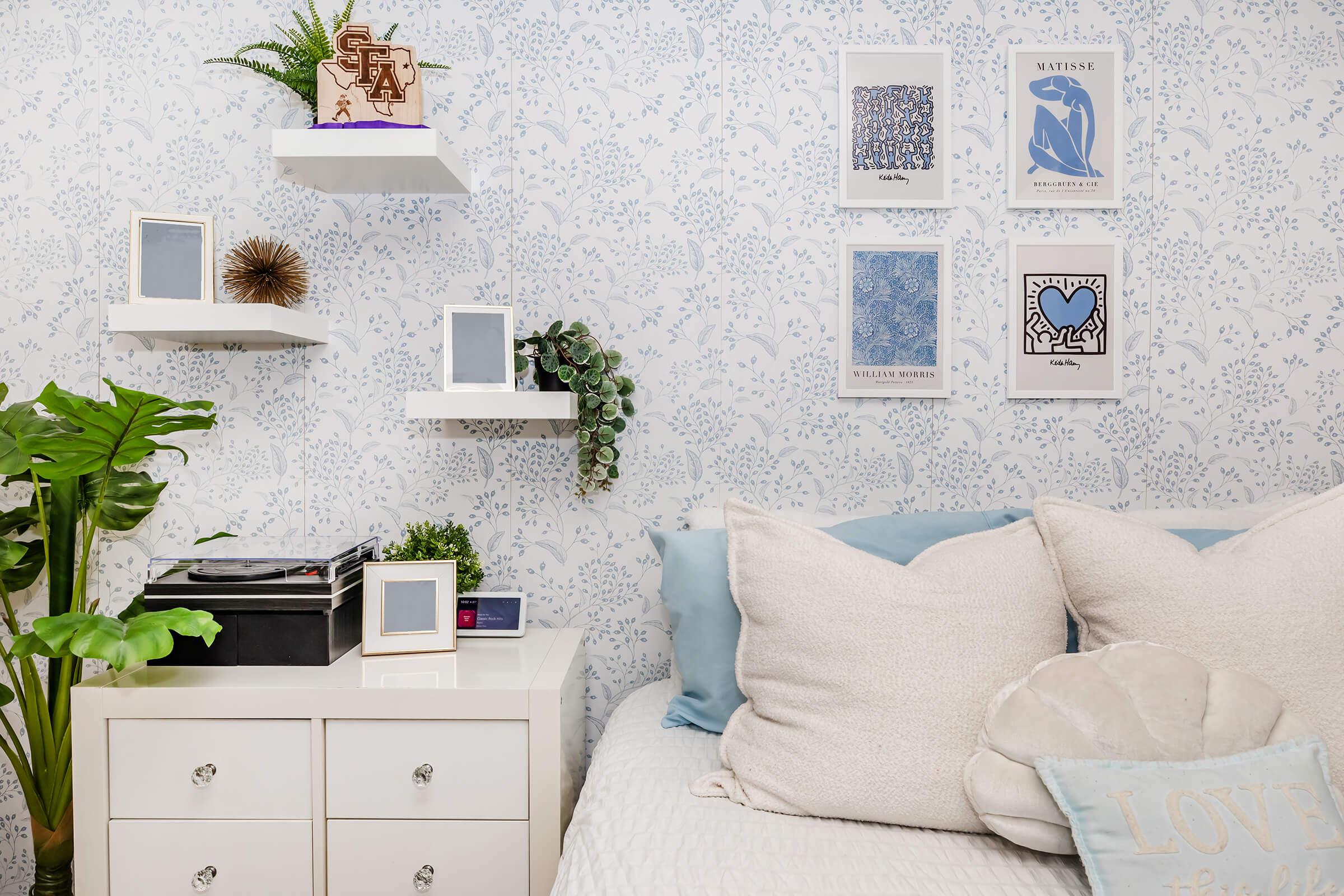 A cozy bedroom corner featuring a white bed with large, soft pillows and a decorative throw pillow. There's a white nightstand with several picture frames and books, surrounded by green plants. The wall is adorned with light blue wallpaper and shelves displaying small decorative items and artwork.