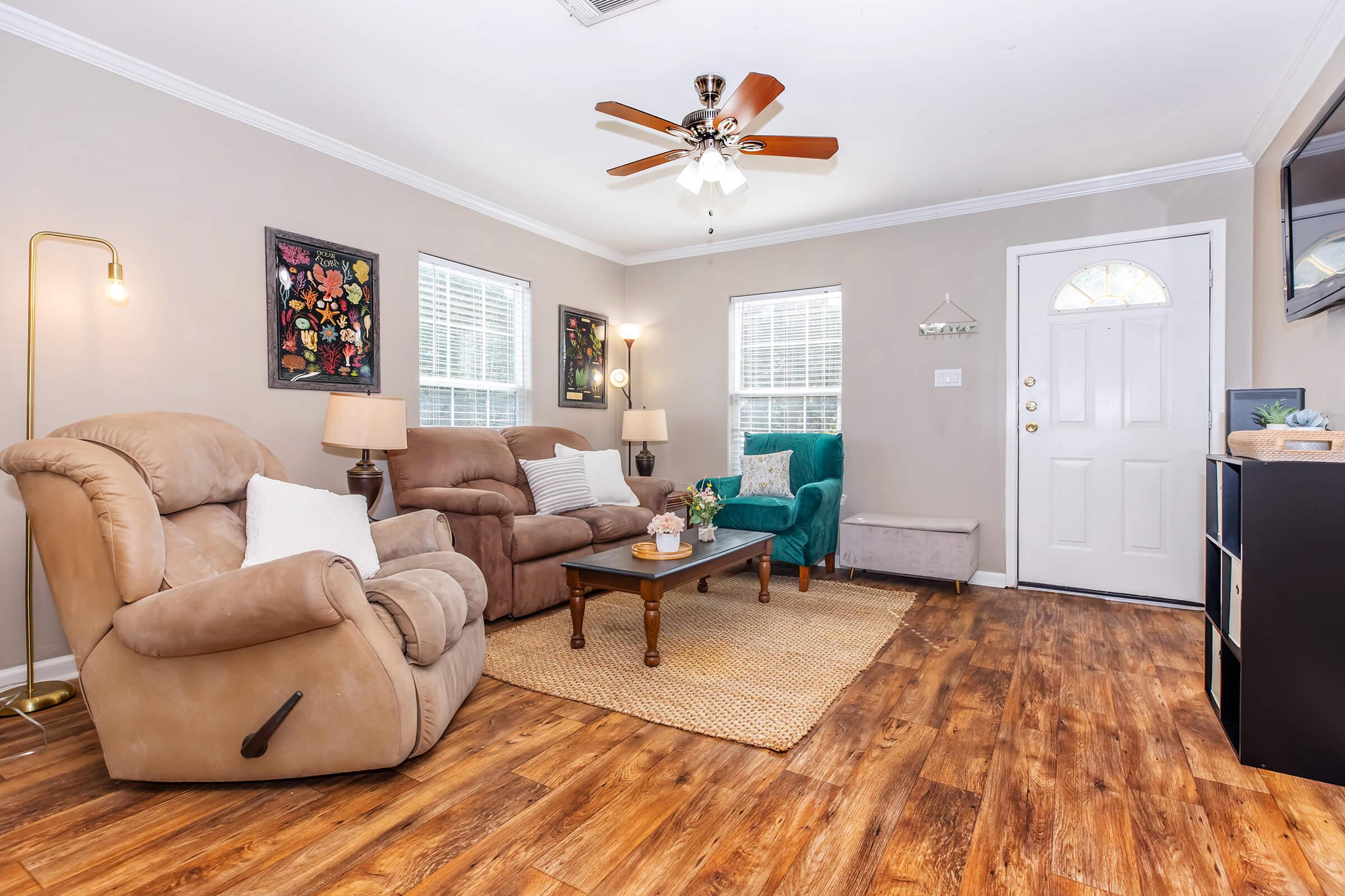 A cozy living room featuring two recliner chairs, a coffee table with a flower vase, and a decorative rug. Light-colored walls and wood-style flooring create a warm atmosphere. There are framed artworks on the walls, two windows allowing natural light, and a door leading outside, enhancing the inviting feel of the space.