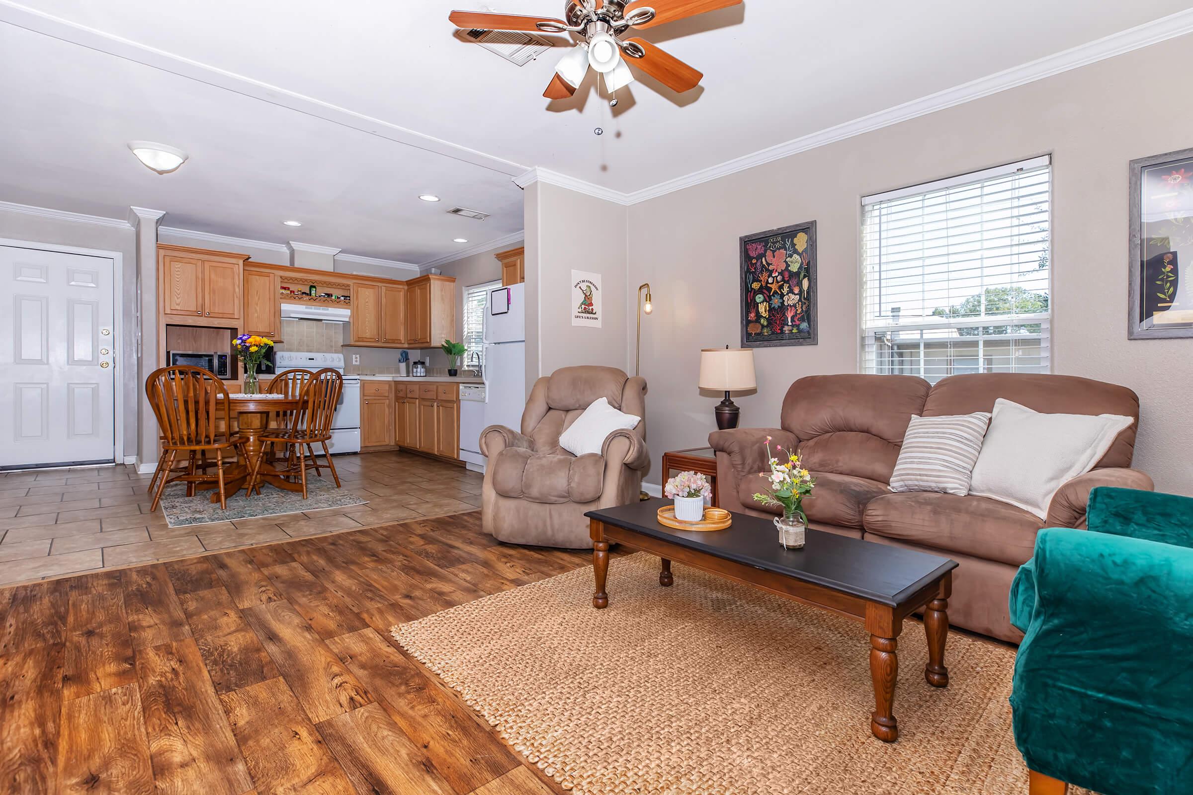 Cozy living room featuring comfortable seating with a coffee table, decorative plants, and a fan overhead. The open kitchen and dining area are visible in the background, showcasing wooden cabinetry and a dining table. Natural light streams in through the windows, creating a warm atmosphere.