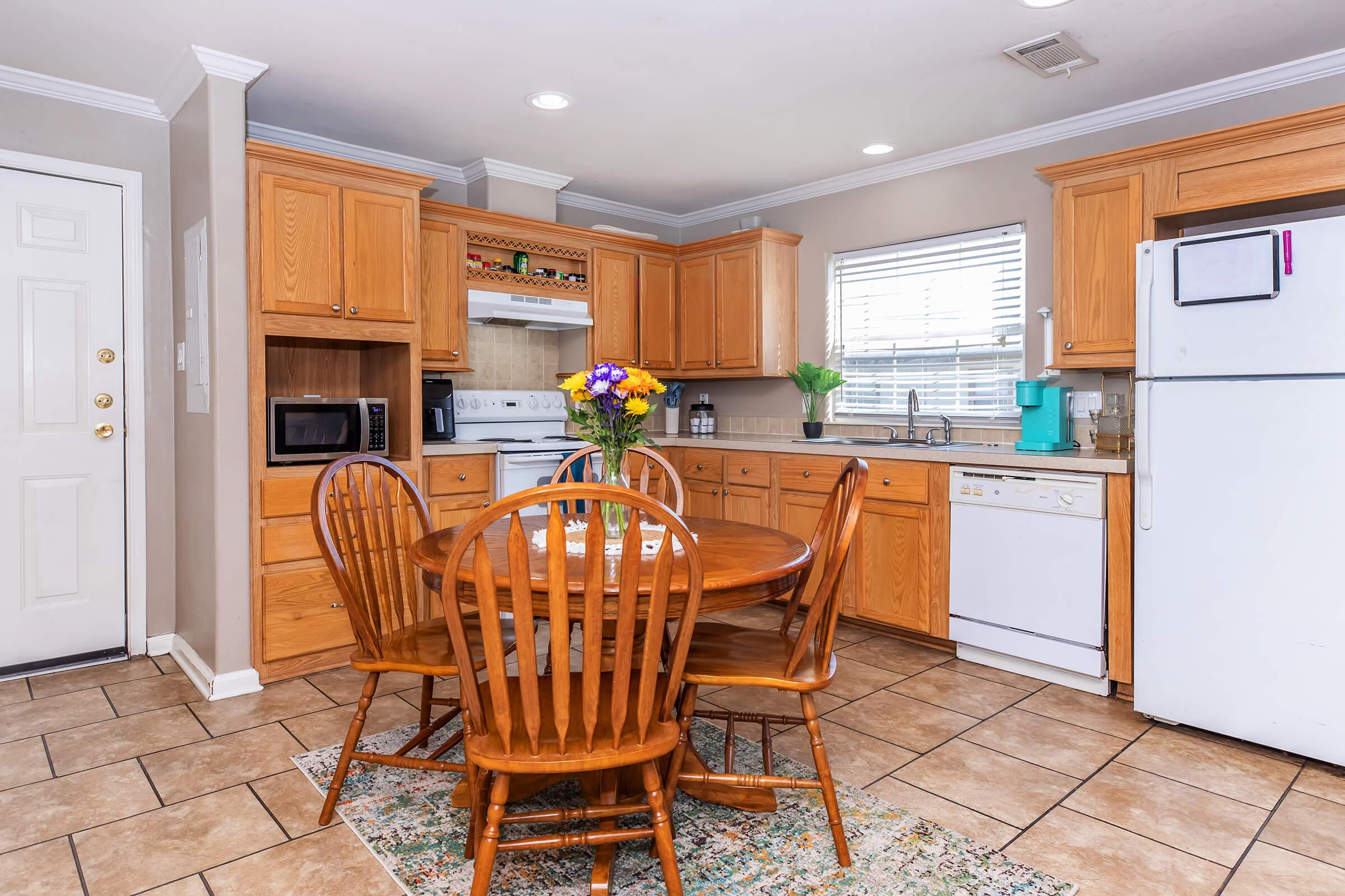 A cozy kitchen with wooden cabinets and a round dining table surrounded by four chairs. The kitchen features essential appliances including a refrigerator, microwave, and dishwasher. A vase of colorful flowers sits on the table, and natural light enters through the window, highlighting the tiled floor.