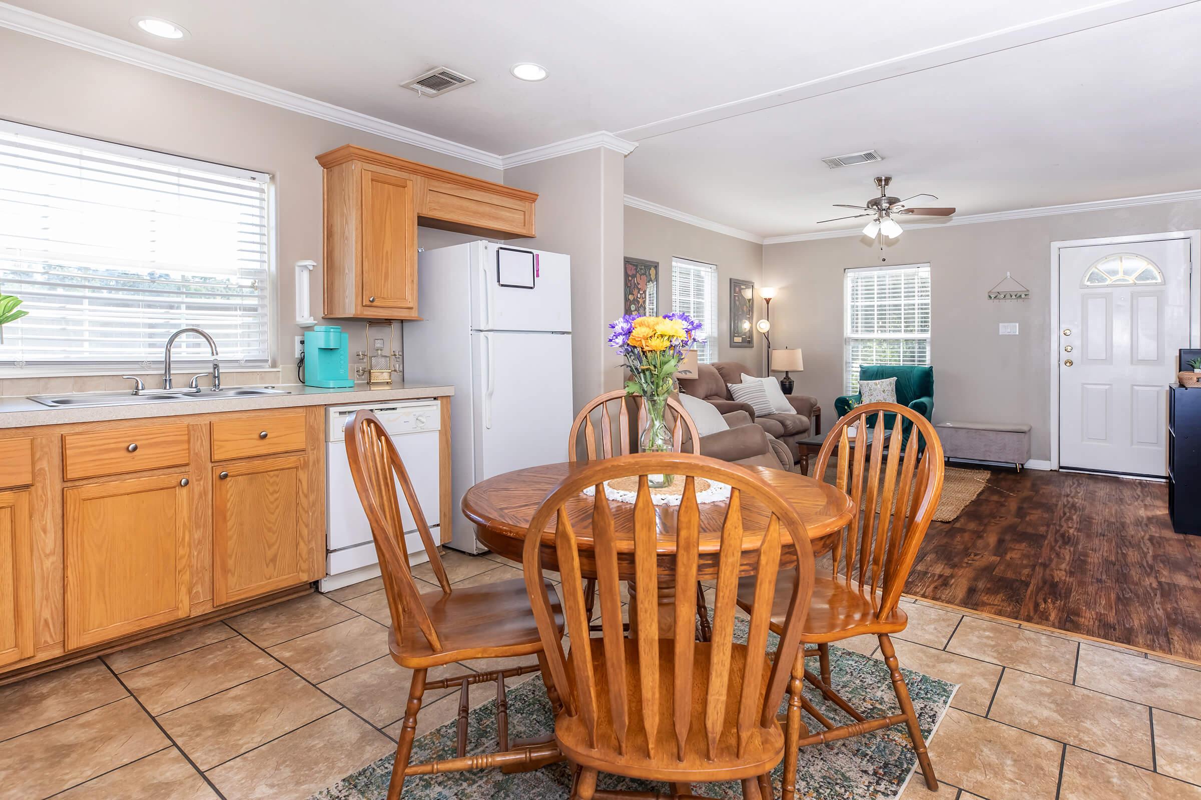 A cozy kitchen and dining area featuring a wooden round table with four chairs, a refrigerator, and a sink. Natural light streams through windows, and a colorful flower vase sits at the center of the table. A living room is visible in the background with a sofa and a ceiling fan, creating an inviting atmosphere.