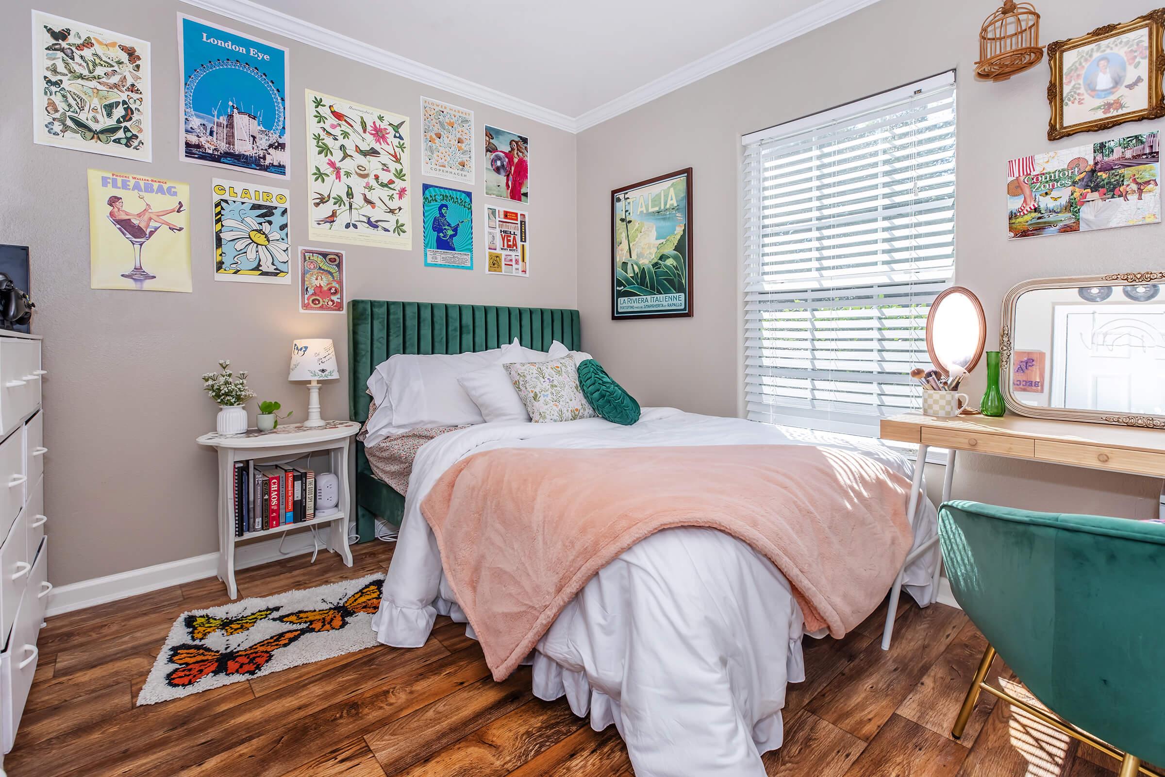 Cozy bedroom featuring a green velvet bed with white bedding and a pink throw blanket. Decorated with various art posters on the walls, a small side table with books, and a desk with a mirror. Sunlight streams in through a window, highlighting the wooden floor and a colorful rug.