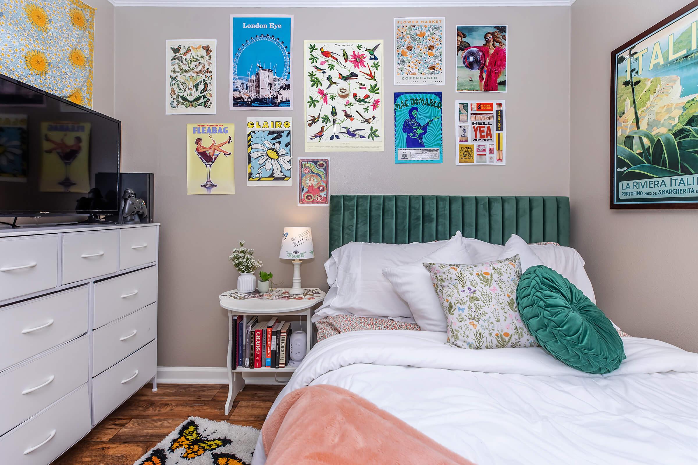 A cozy bedroom featuring a white bed adorned with patterned pillows, a green upholstered headboard, and a white duvet. A nightstand with a lamp sits beside the bed, while a dresser with a TV is nearby. The walls are decorated with colorful art prints, including travel posters and floral designs. A soft rug adds warmth to the wooden floor.