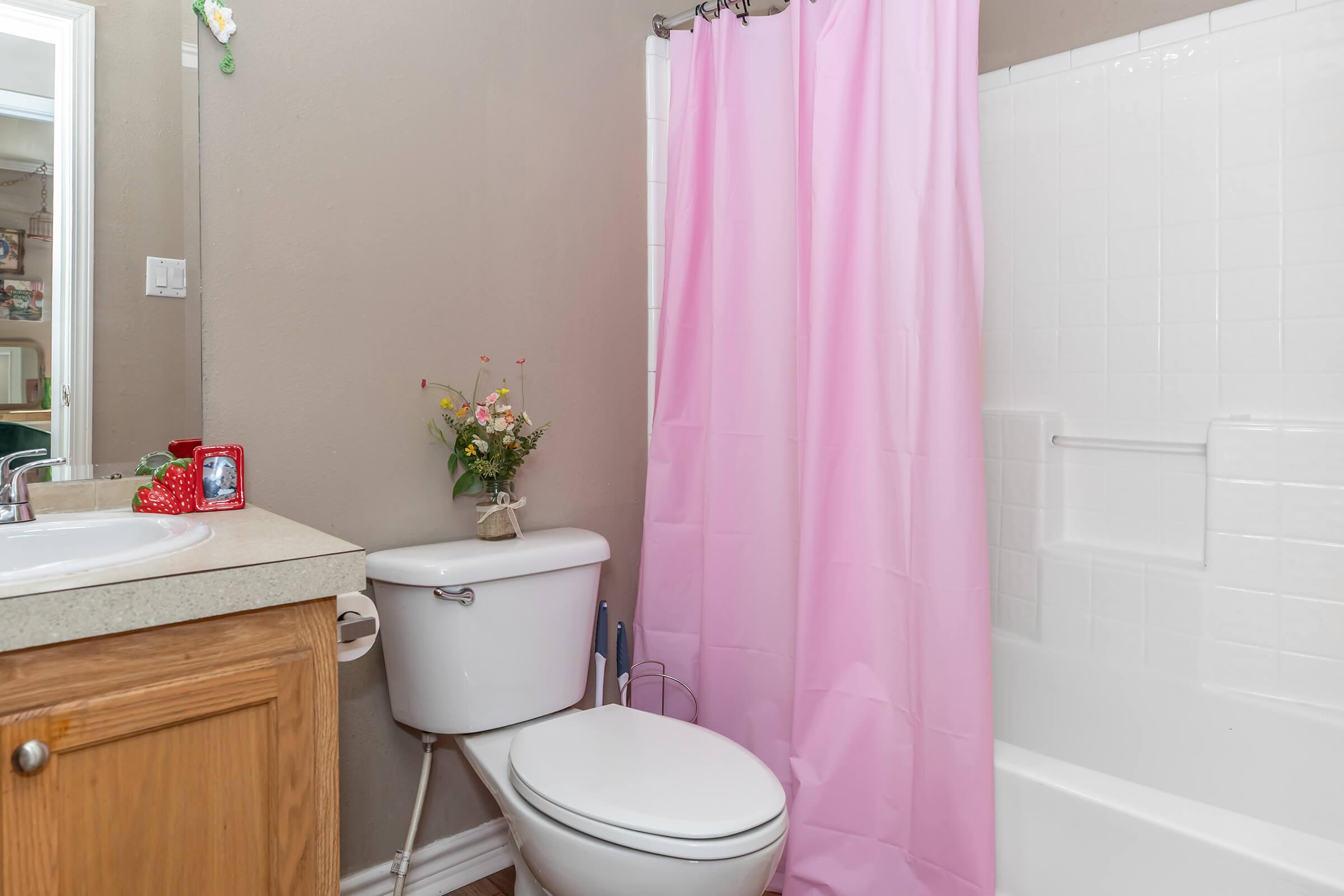 A clean bathroom with a light pink shower curtain, a white toilet, and a wooden vanity. A small vase with flowers sits on the vanity. The bathroom features white tiled walls and a bathtub with a showerhead, creating a bright and inviting atmosphere.