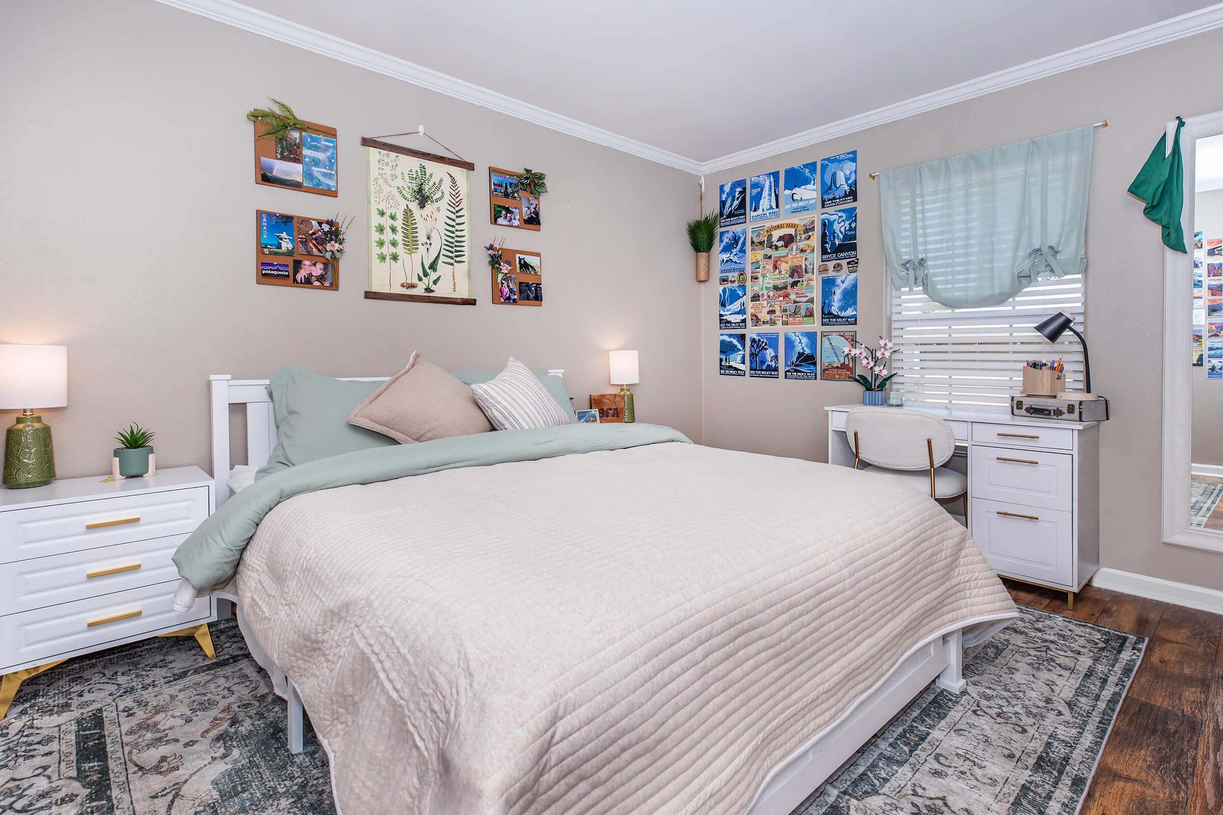 A cozy bedroom featuring a white bed with soft bedding, decorative pillows, and ambient lighting. The walls are adorned with framed nature-themed artwork and photo collages. A small desk with a chair is visible, along with a green curtain and indoor plants, creating a warm and inviting atmosphere.