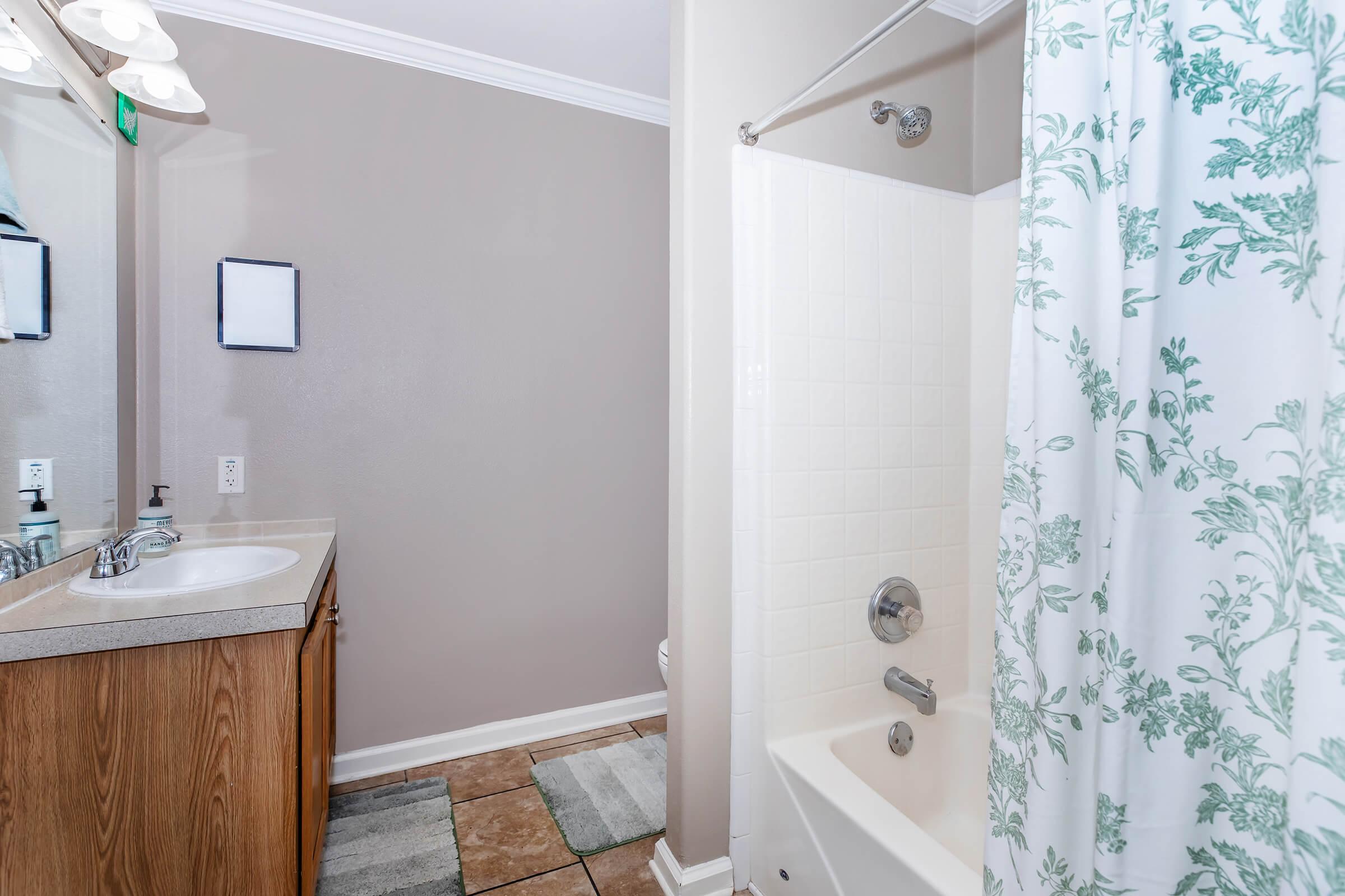 A well-lit bathroom featuring a bathtub with a shower curtain adorned with floral patterns. The countertop has a sink and a mirror, with wooden cabinetry below. The floor is tiled, and there's a small area rug near the bathtub. The walls are painted a neutral color, creating a clean and inviting atmosphere.