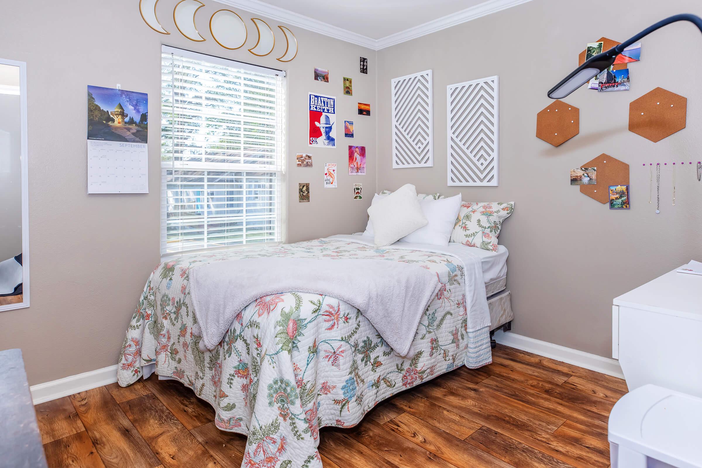 Cozy bedroom with a neatly made bed featuring a floral bedspread. Walls adorned with decorative items, including photos and wall art. A window with curtains lets in natural light, and a small desk with a lamp is visible. The floor has warm wooden tones, adding to the inviting atmosphere.