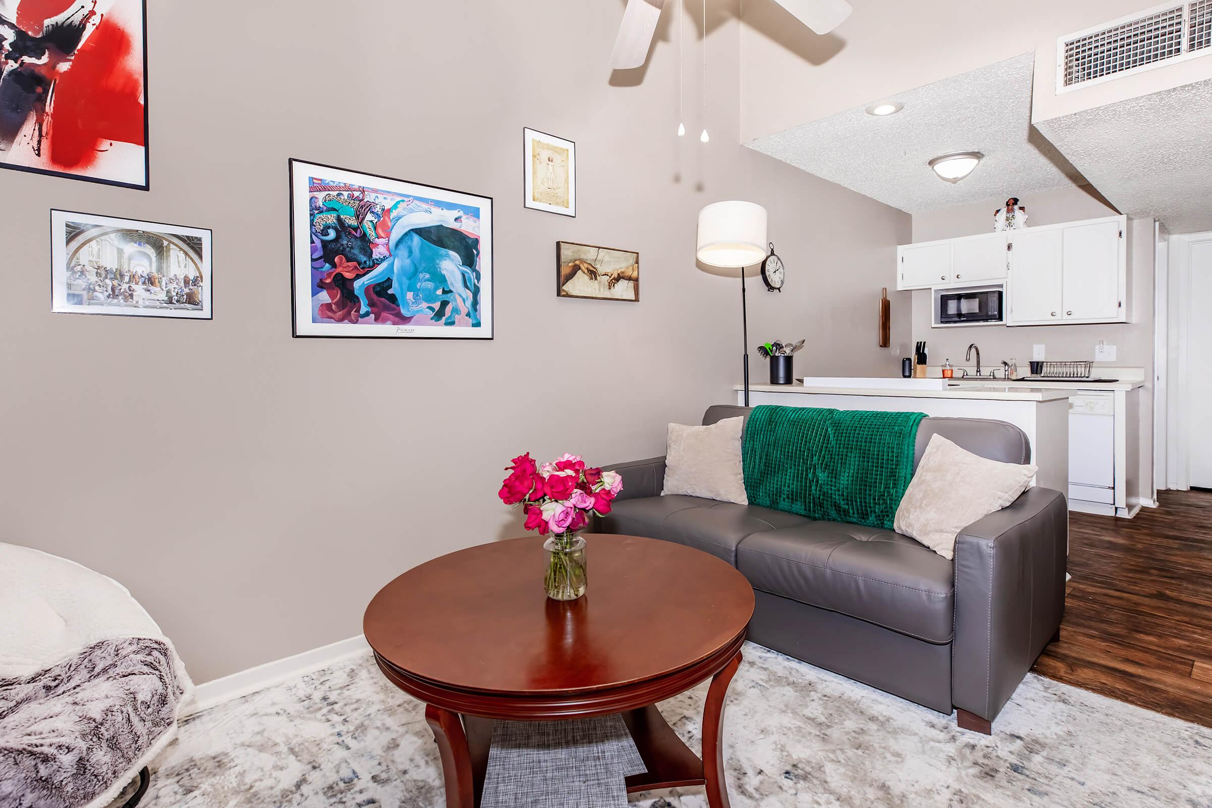 A cozy living room featuring a gray sofa with a green throw blanket, a round wooden coffee table with a vase of pink flowers, and various framed artwork on the walls. In the background, a kitchen area is visible with white cabinetry and appliances. The space has warm lighting and wooden flooring.