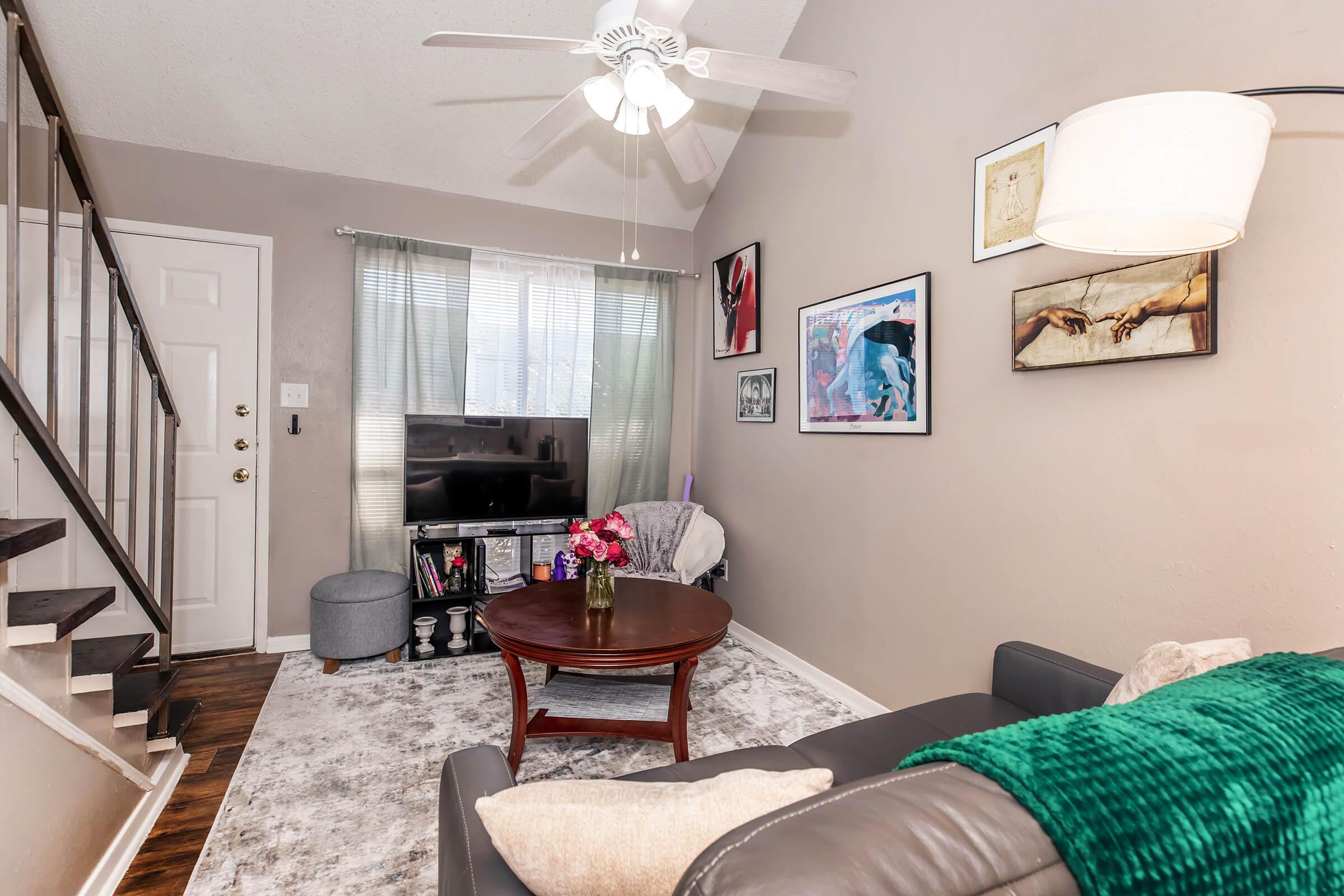 A cozy living room featuring a gray sofa with green throw, a round wooden coffee table adorned with flowers, and a TV stand. The room has beige walls, light curtains, and artwork on display. A ceiling fan and a staircase lead to an upper level, creating a warm and inviting atmosphere.